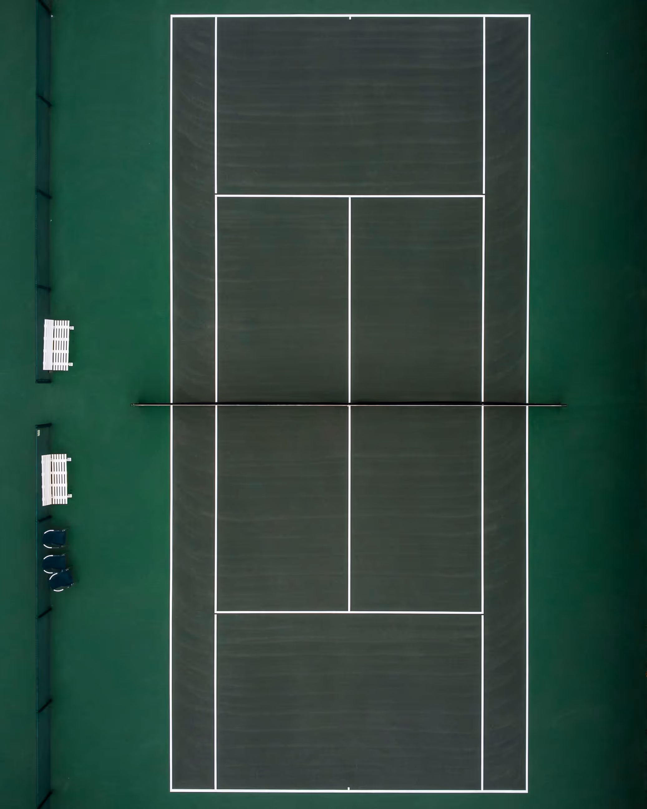 Eine Vogelperspektive eines professionellen Tennisplatzes mit grüner Oberfläche und weißen Linien. An der Seite stehen zwei Tennisbänke und einige blaue Stühle.