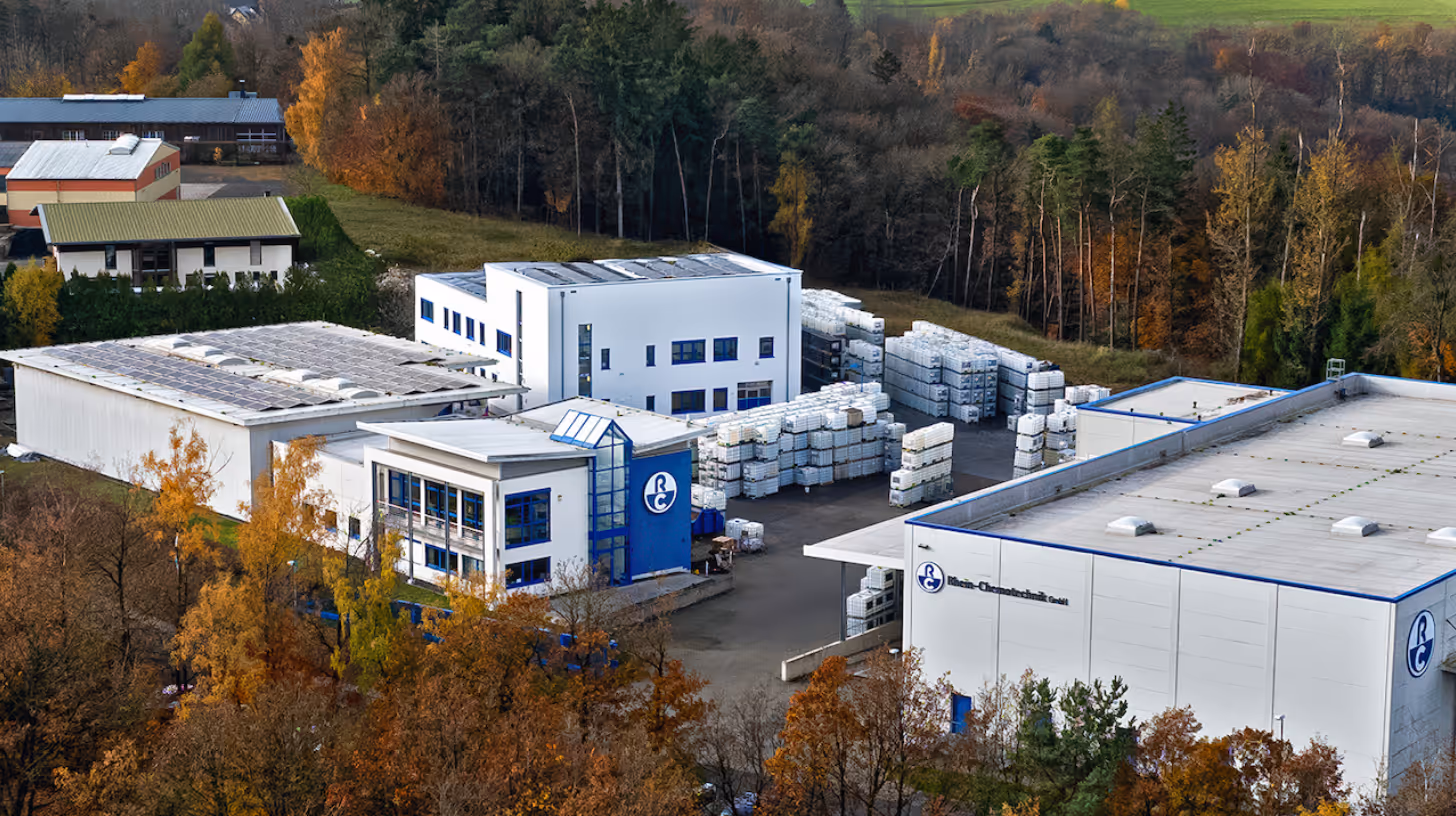 Eine Luftaufnahme eines modernen Industriegebäudes mit weißen Wänden und blauen Akzenten, umgeben von buntem Herbstlaub. Im Hintergrund sind weitere Gebäude sichtbar, die in die grüne Landschaft integriert sind.