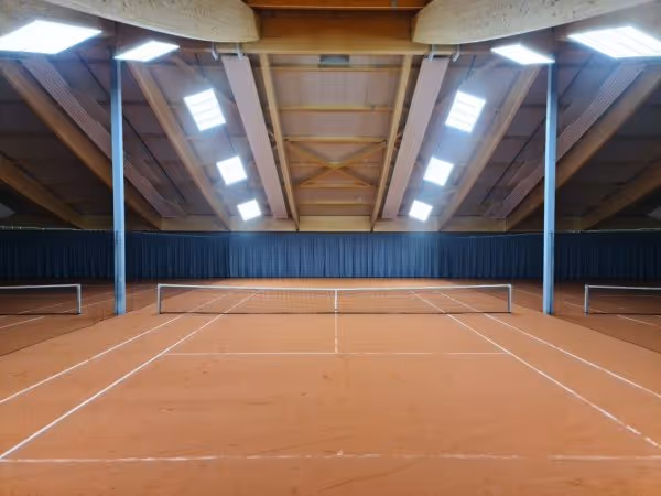 Eine Innenansicht einer Tennisanlage mit zwei Spielfeldern auf rotem Sand. Die hohen Decken sind mit Holzstäben und Tageslichtlampen ausgestattet, was eine helle und offene Atmosphäre schafft.