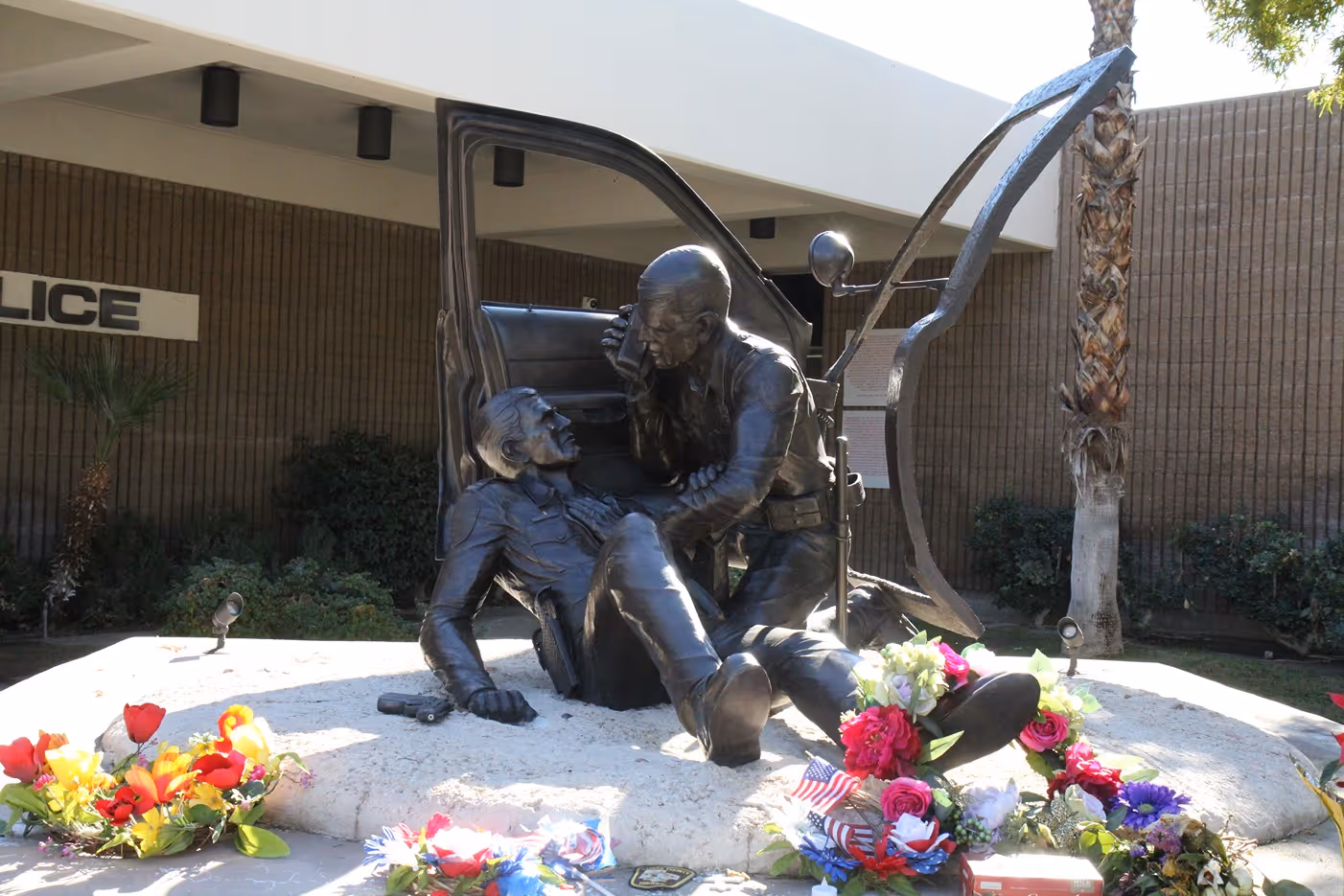 A bronze sculpture depicts a police officer assisting an injured colleague, surrounded by flowers and mementos at a police station.