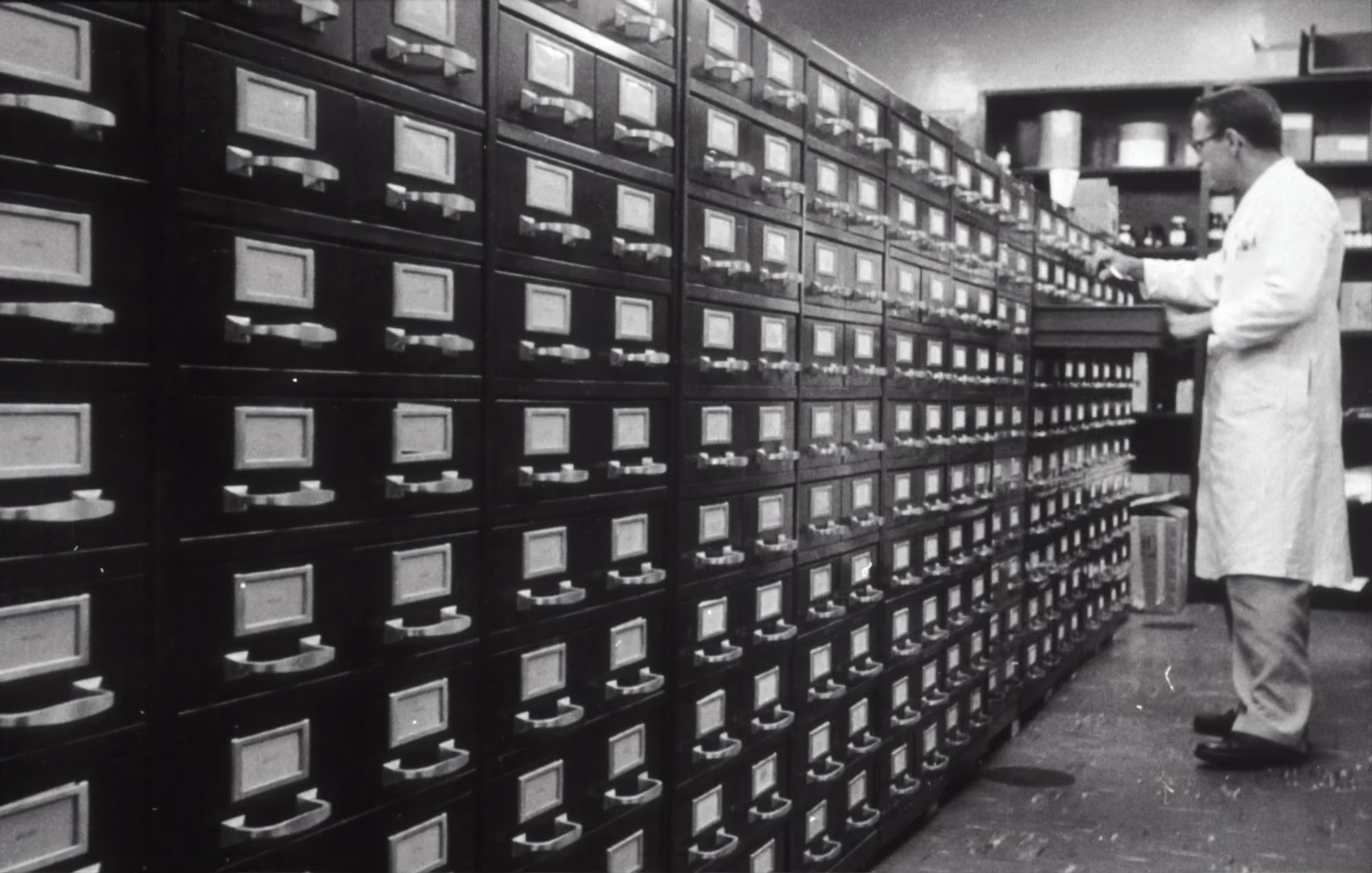 Man in a white lab coat organizing files in a large wall of metal filing drawers.