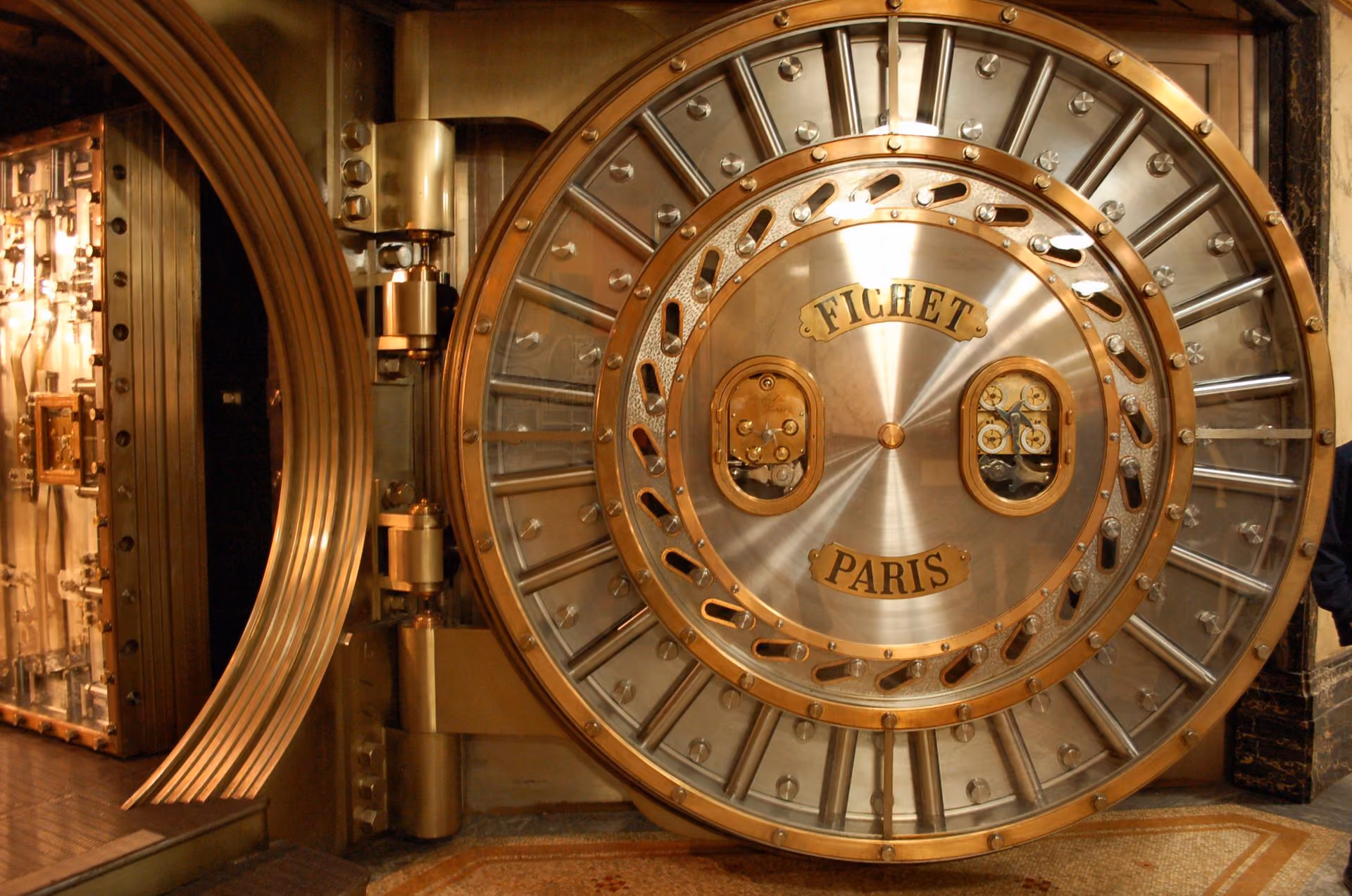 Large open round bank vault door with metal bolts and dials labeled Fichet Paris.