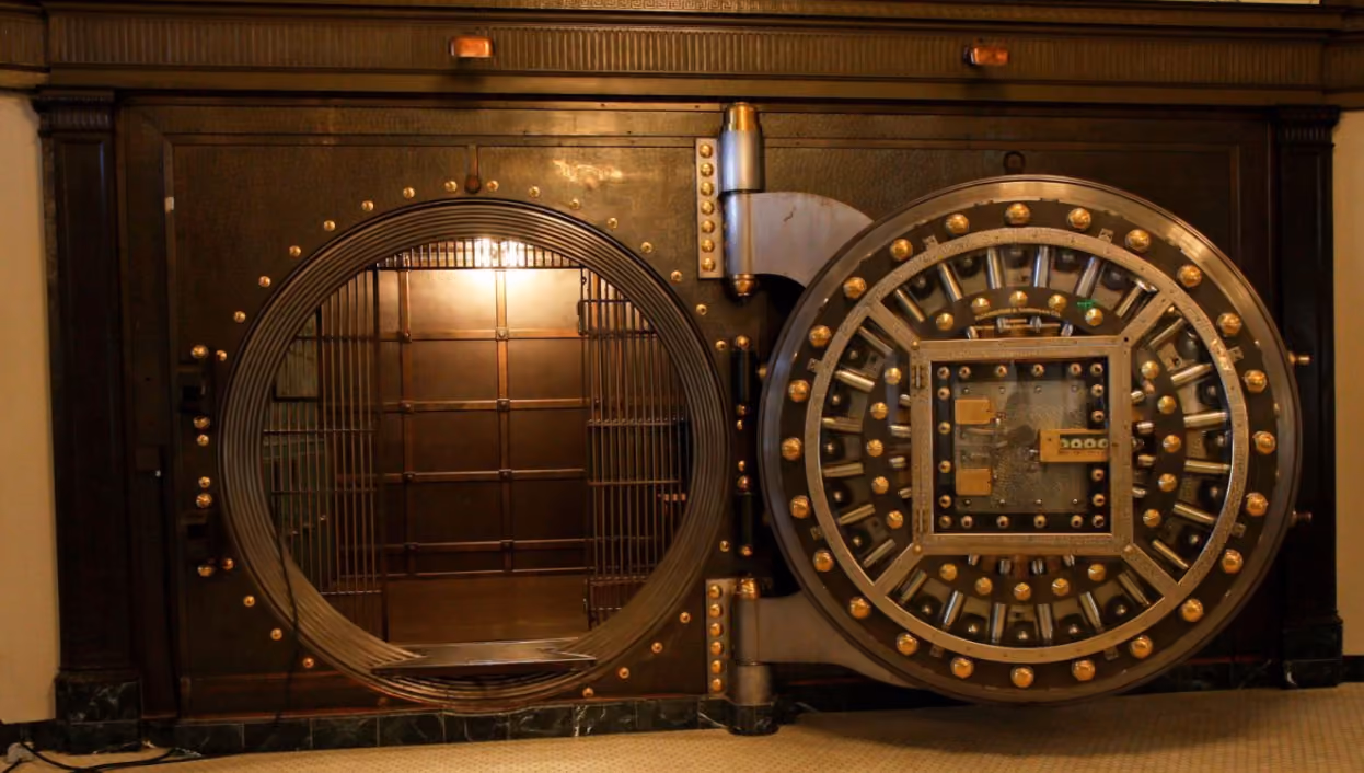 Open vintage bank vault door with circular locking mechanism and metal bars inside room