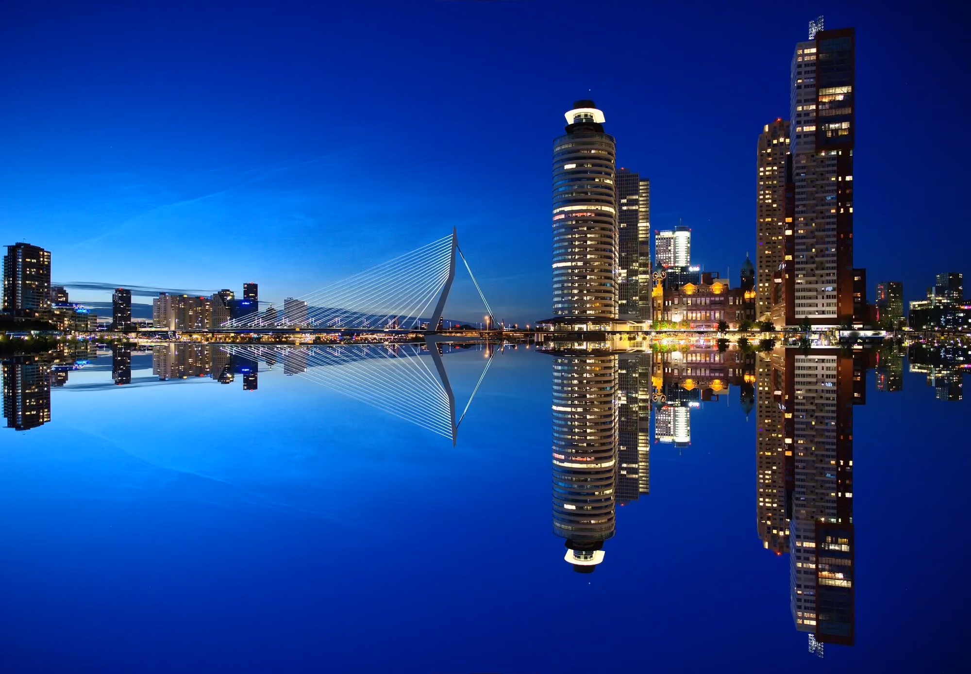 City skyline at dusk with illuminated buildings and a bridge reflected in calm water.