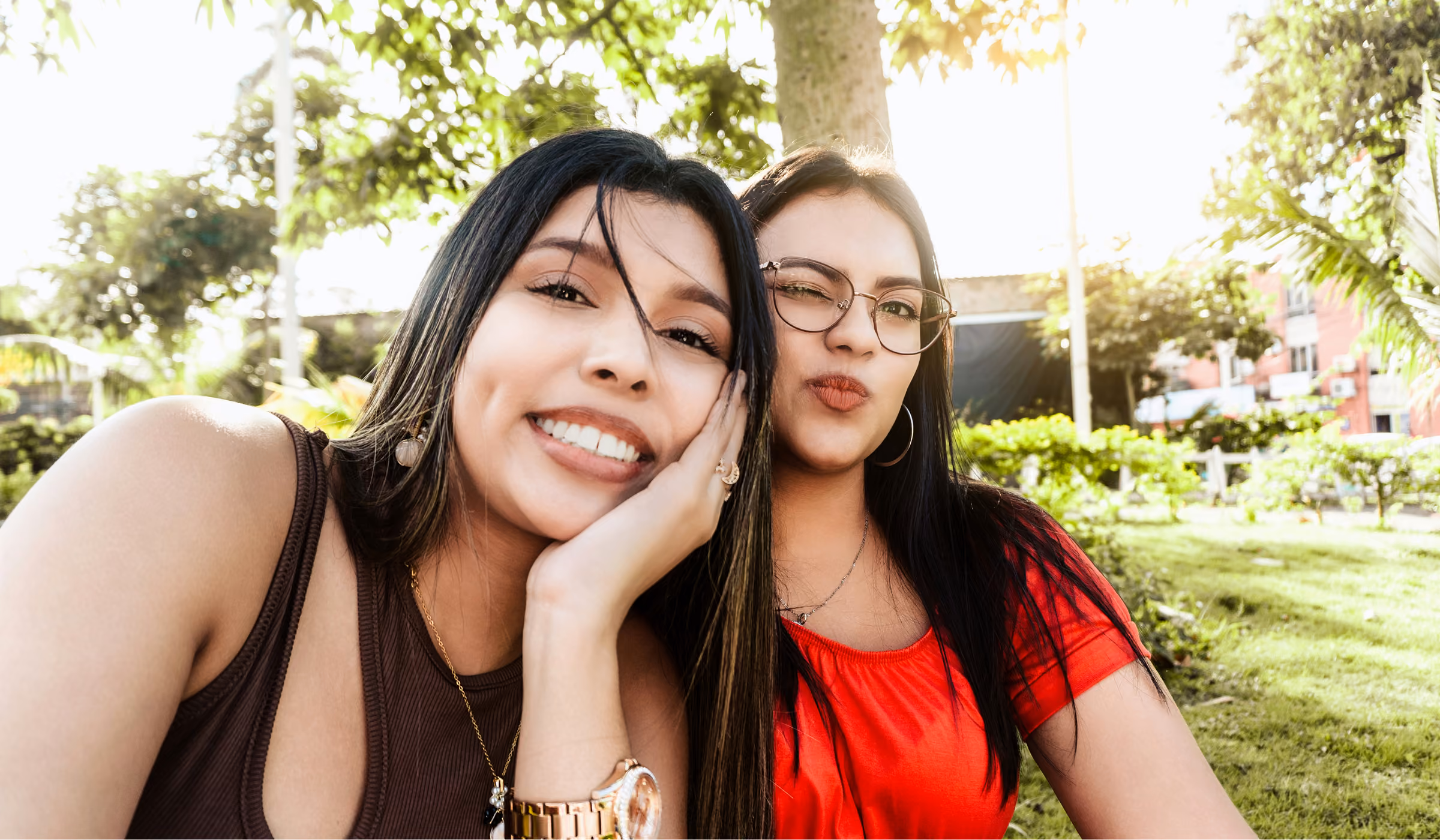 Two smiling young women posing closely outdoors with trees and buildings in the background.