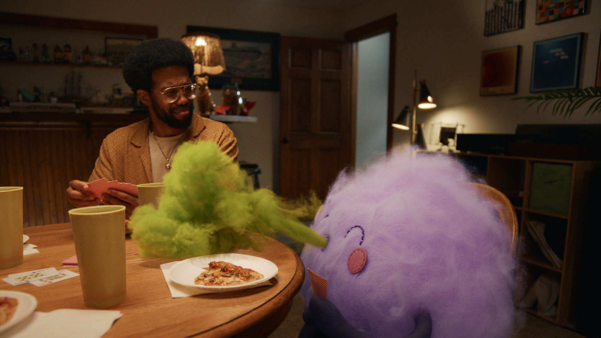 A man with glasses and an afro looks confused while sitting at a table playing cards with a fluffy purple creature that exhales a cloud of green vapor toward a slice of pizza on a paper plate.