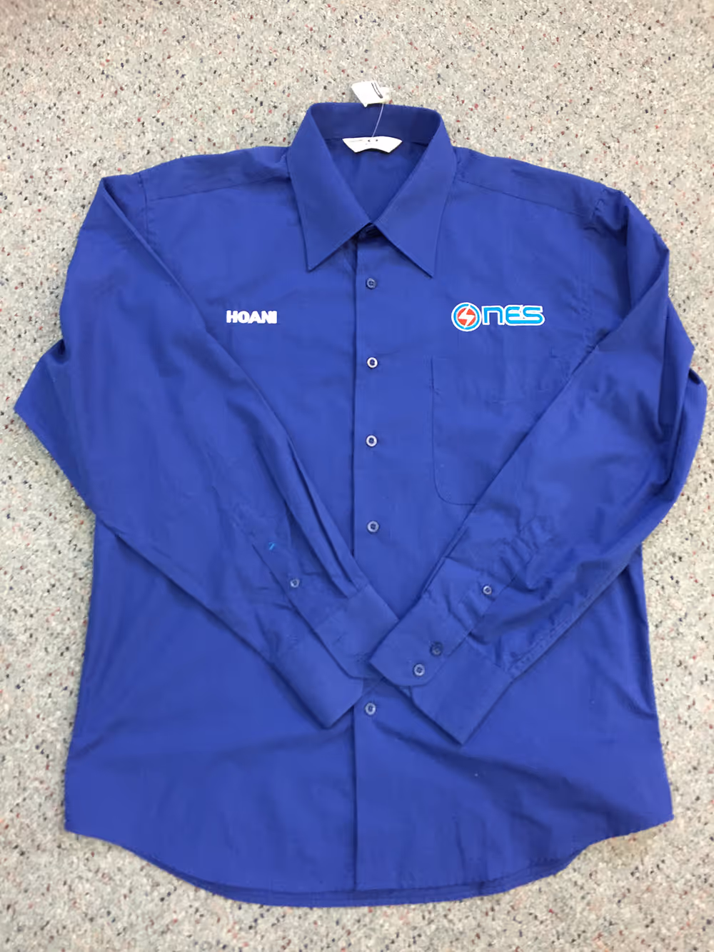A dark blue shirt with the NES logo embroidered on one side and the staff members name embroidered on the other side