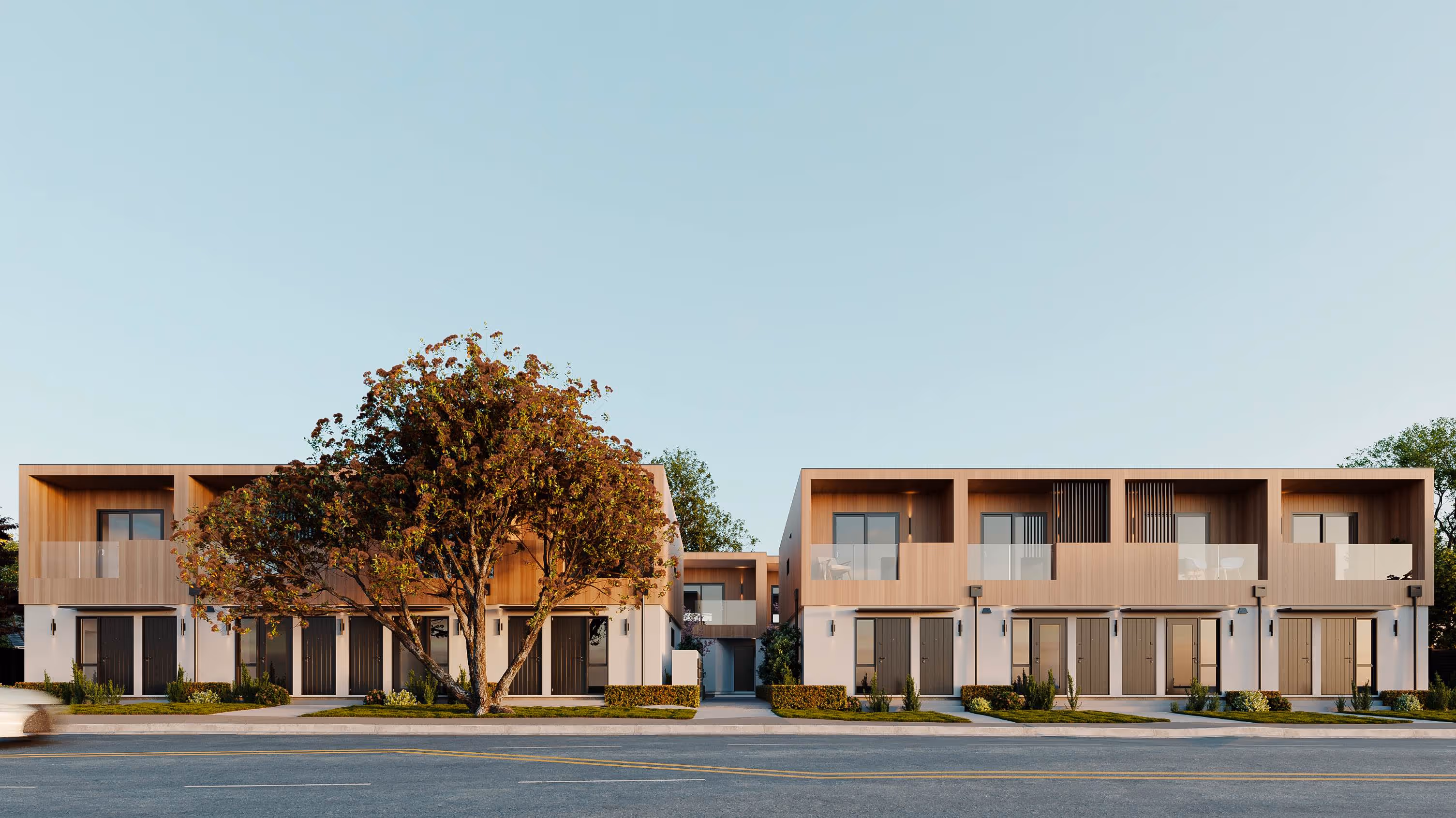 Modern residential townhouse development with warm wood and white stucco facades, featuring two-story units with private balconies, mature landscaping including a prominent autumn tree, viewed from across a paved street