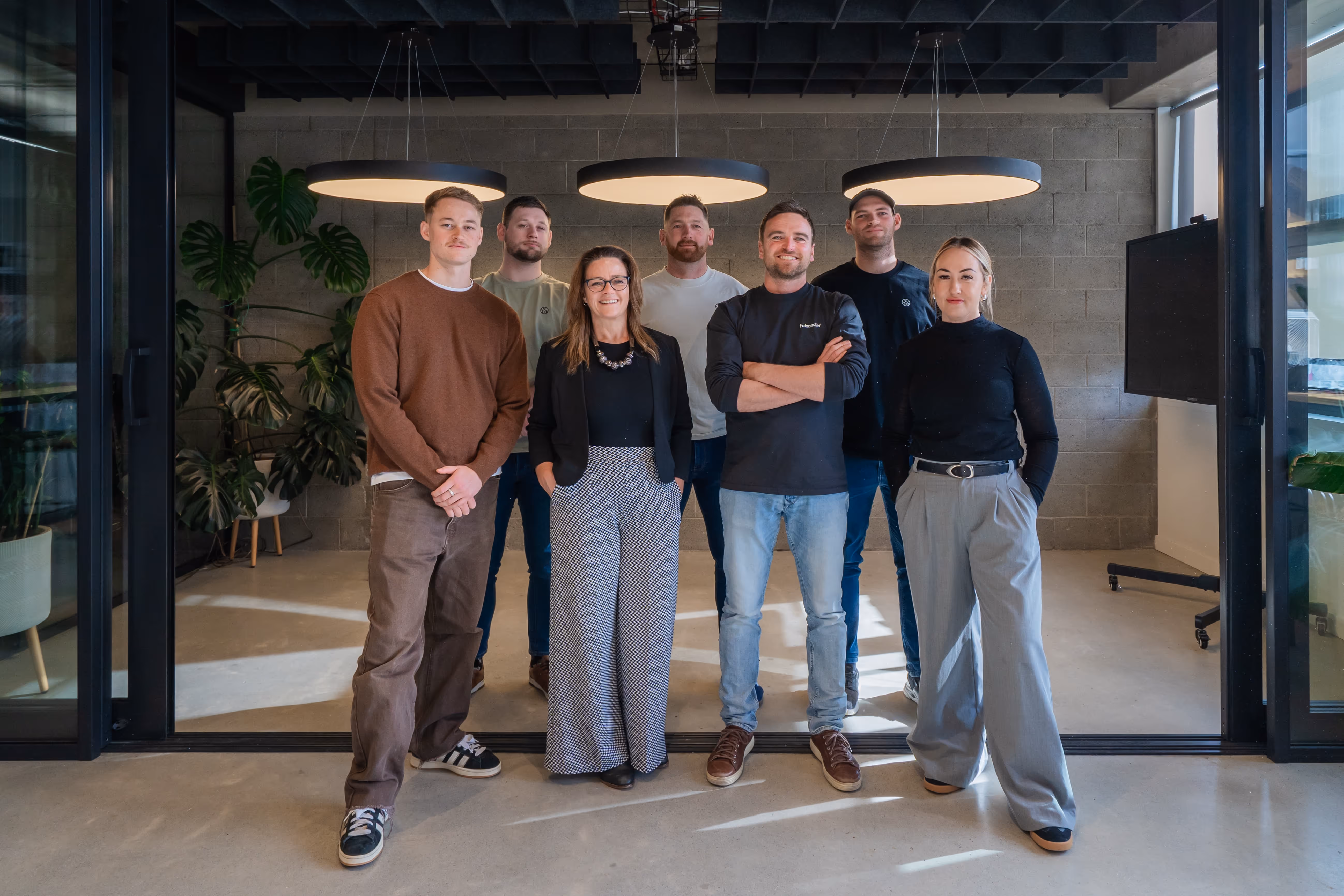 Seven members of the Faisandier team standing together in modern industrial office space with exposed concrete block wall, three circular pendant lights overhead, polished concrete floors, glass partitions, and indoor plants, dressed in smart casual business attire.