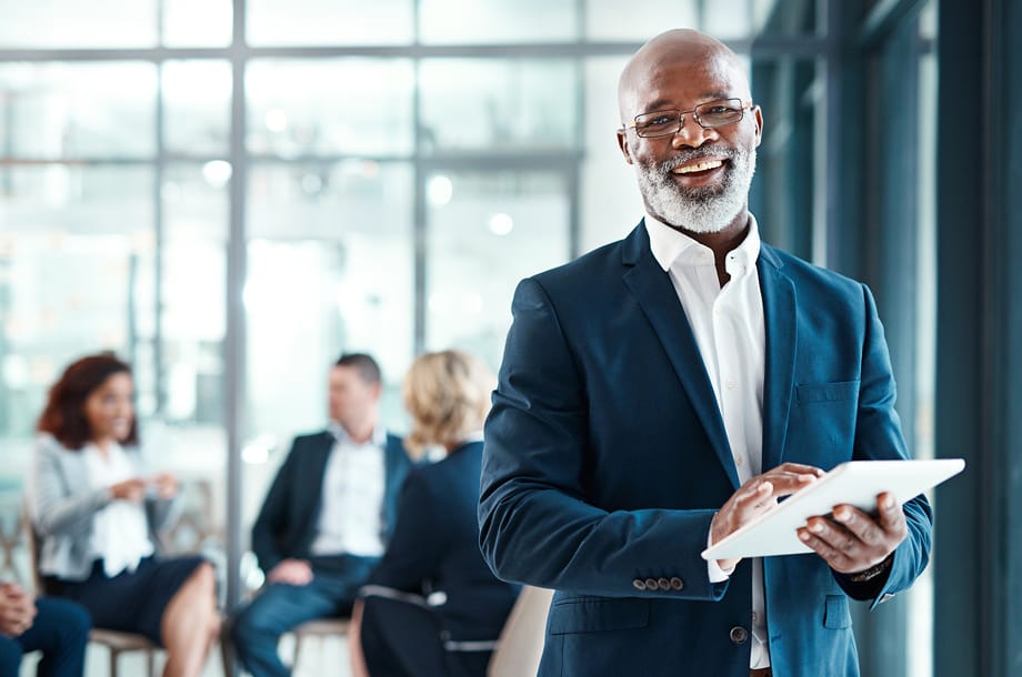 Smiling businessman in glasses and suit using a digital tablet in a modern office with colleagues in the background.
