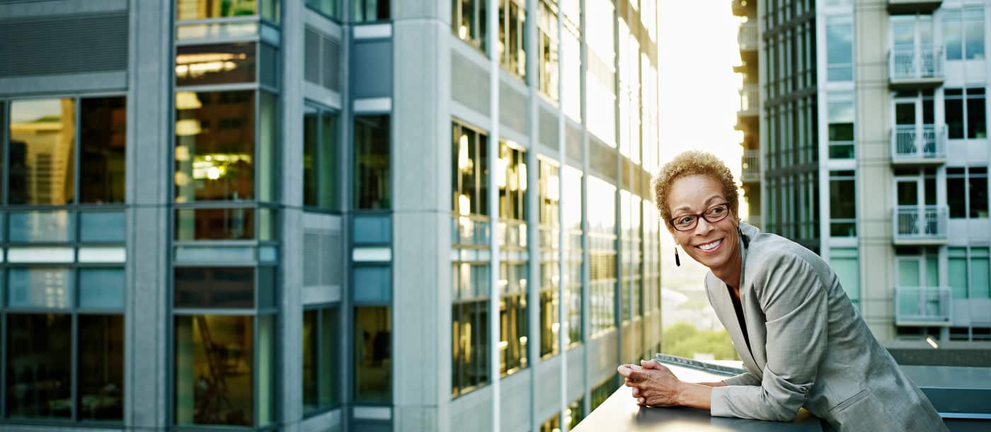 Smiling professional woman with short curly hair and glasses leaning on a railing outside modern office buildings.