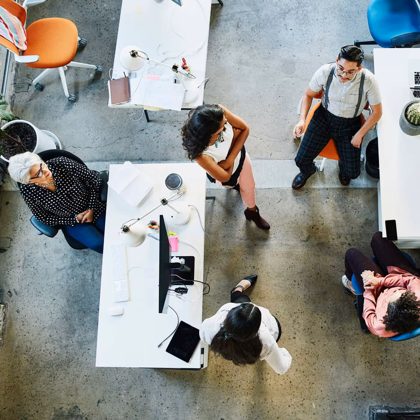 Overhead view of five diverse coworkers engaged in discussion around white office desks in a modern workspace.