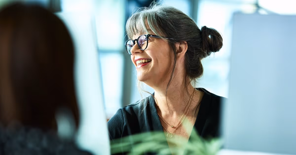 Smiling mature woman with gray hair and glasses in a black top, sitting indoors near a computer screen.