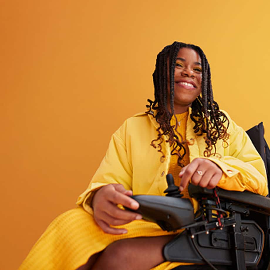 Smiling woman with braided hair wearing yellow clothes sitting in an electric wheelchair against a yellow background.