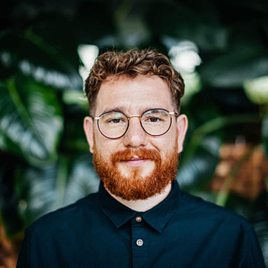 Bearded man with curly hair and round glasses wearing a dark button-up shirt, standing in front of leafy green plants.