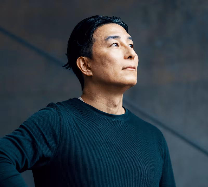 Portrait of a man with short black hair and a serious expression, wearing a dark long-sleeve shirt