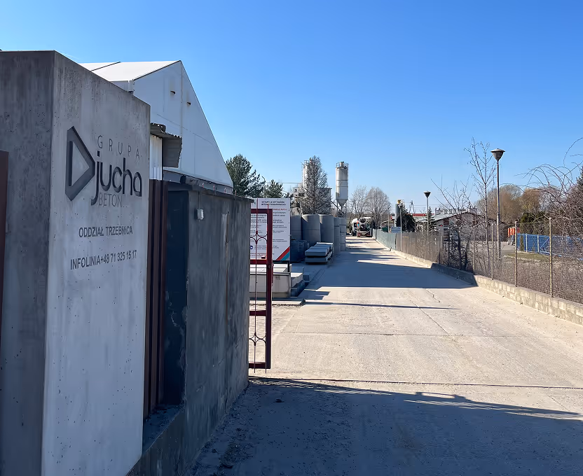 Wejście do zakładu betoniarskiego Grupa Jucha Beton w Trzebnicy, widok na betonowe elementy i silosy na tle niebieskiego nieba.