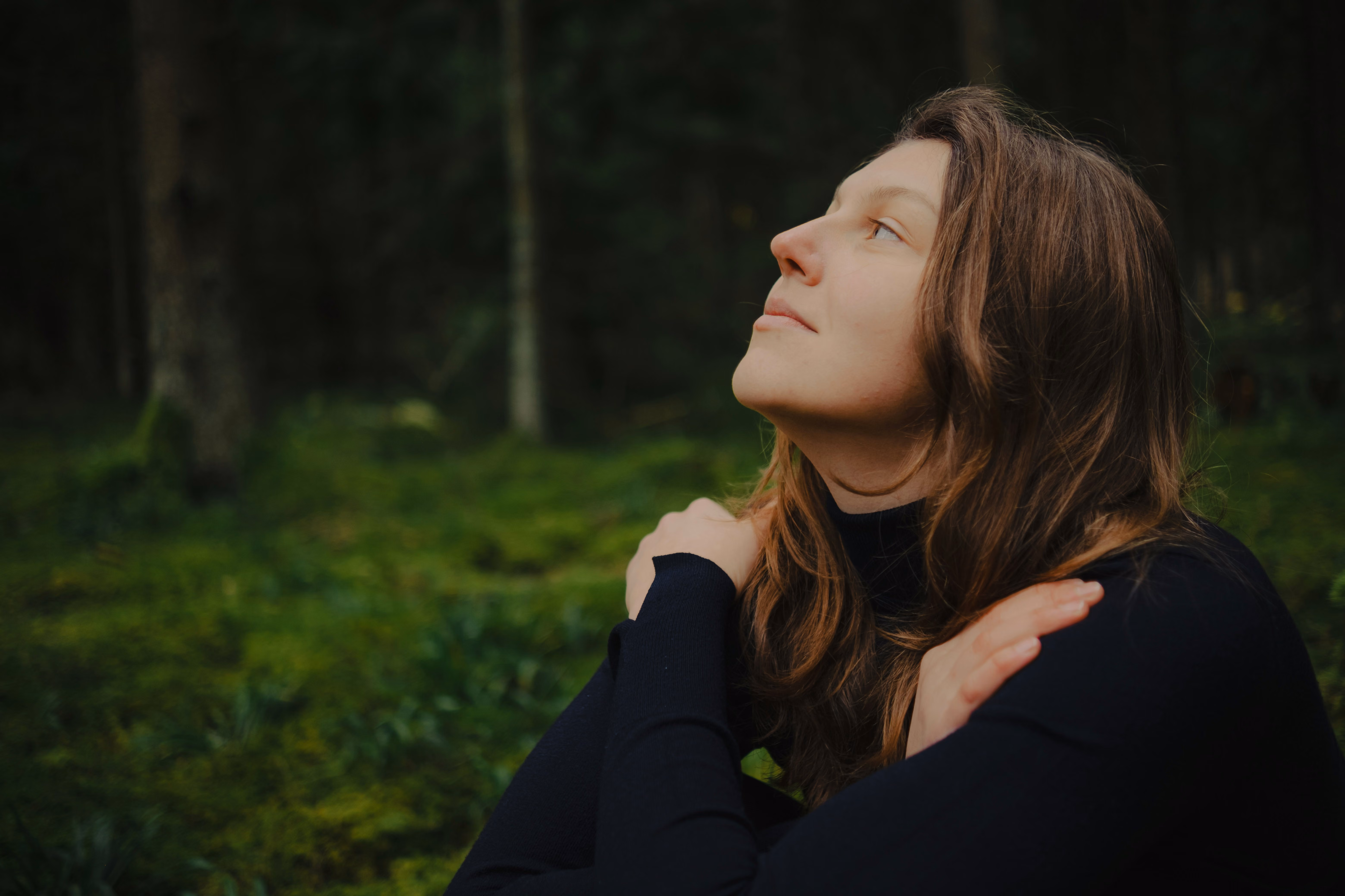 A woman sitting peacefully in a forest, looking upward with her eyes closed
