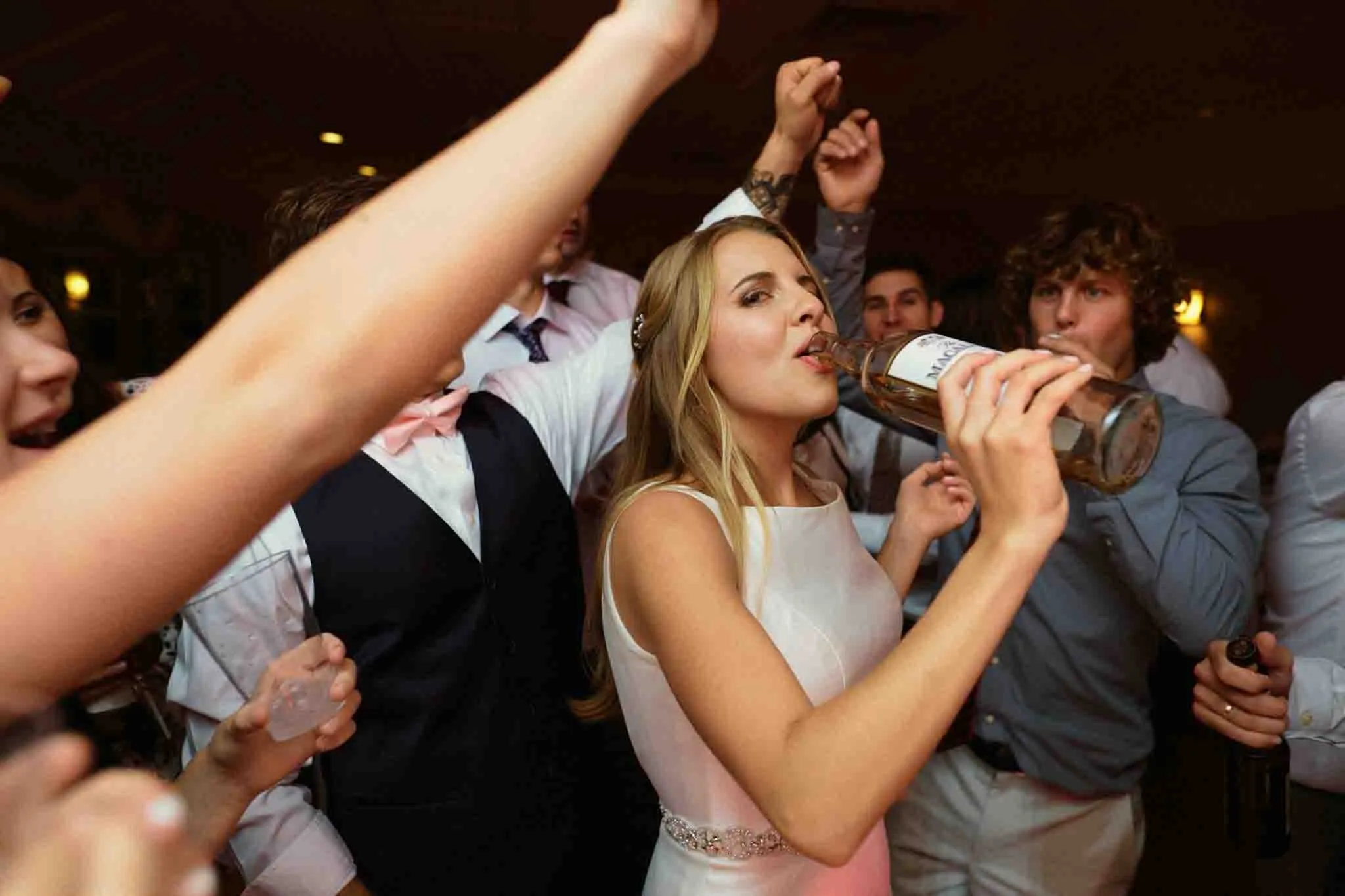 A bride in a white dress sips from a bottle, surrounded by friends with raised arms and bright smiles at a lively indoor party—captured in pure candid film wedding photos New Jersey style.
