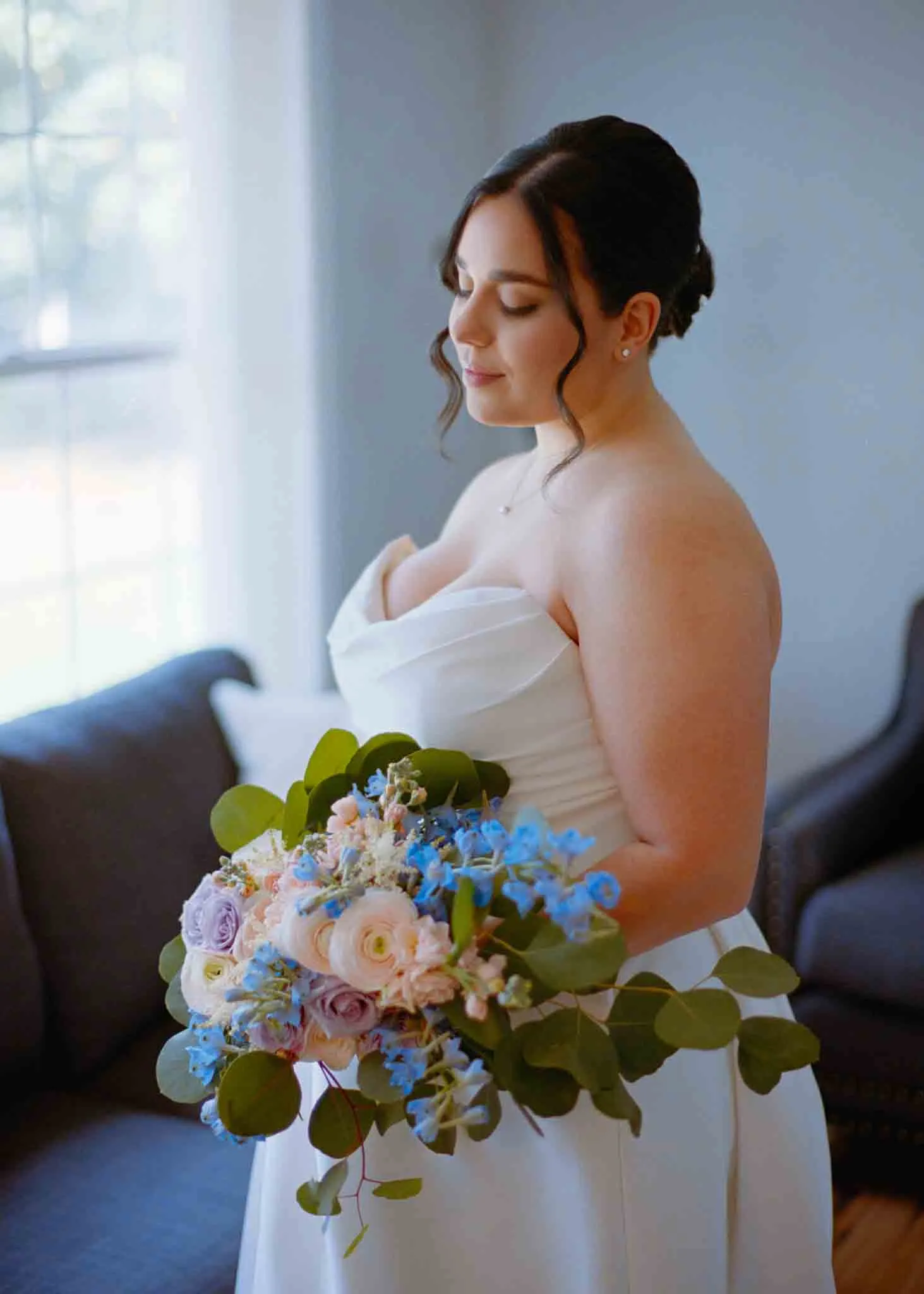 A bride in a white strapless gown holds a bouquet of pastel flowers and greenery, standing indoors by a window as soft natural light illuminates her face—an elegant moment captured by a film wedding photographer NJ.