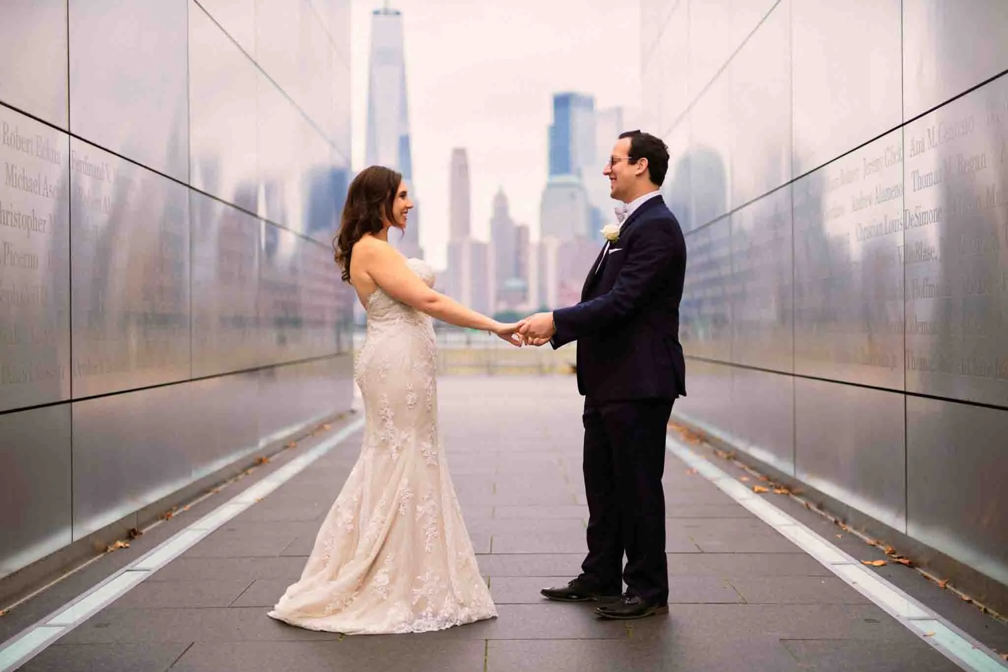 A bride and groom stand facing each other, holding hands in an outdoor urban setting with tall buildings behind them. Captured by a New Jersey film wedding photography expert, their formal attire shines amid reflective city walls.