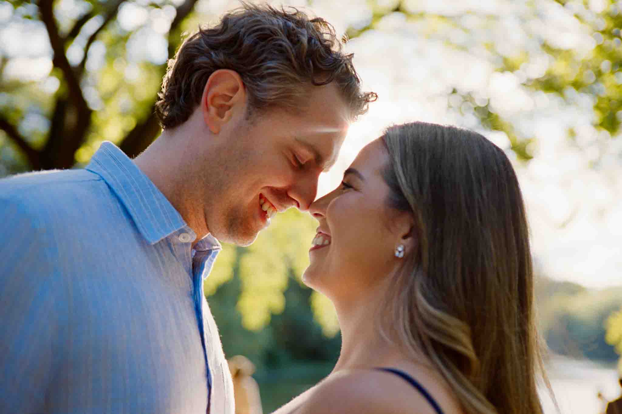 A couple stands close together outdoors, smiling and touching noses as sunlight filters through blurred green trees—a perfect moment captured by a New Jersey film wedding photographer.