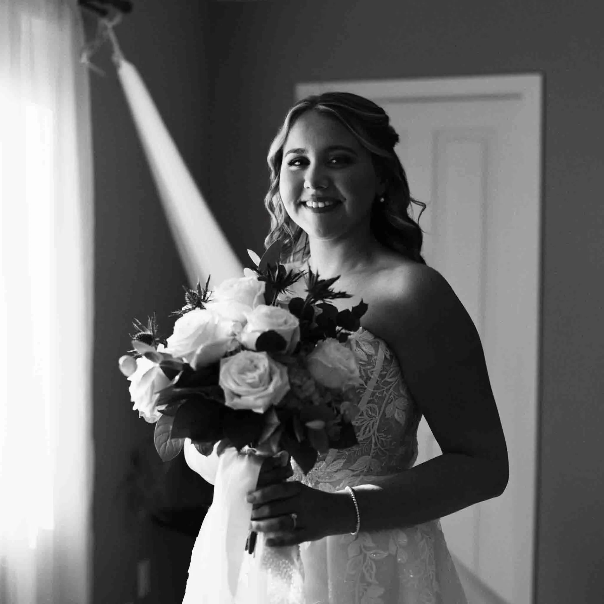 A bride in a strapless lace wedding gown holds a bouquet of roses and greenery, smiling by a sunlit window. Captured in black and white, this moment reflects the timeless style of New Jersey film wedding photography.