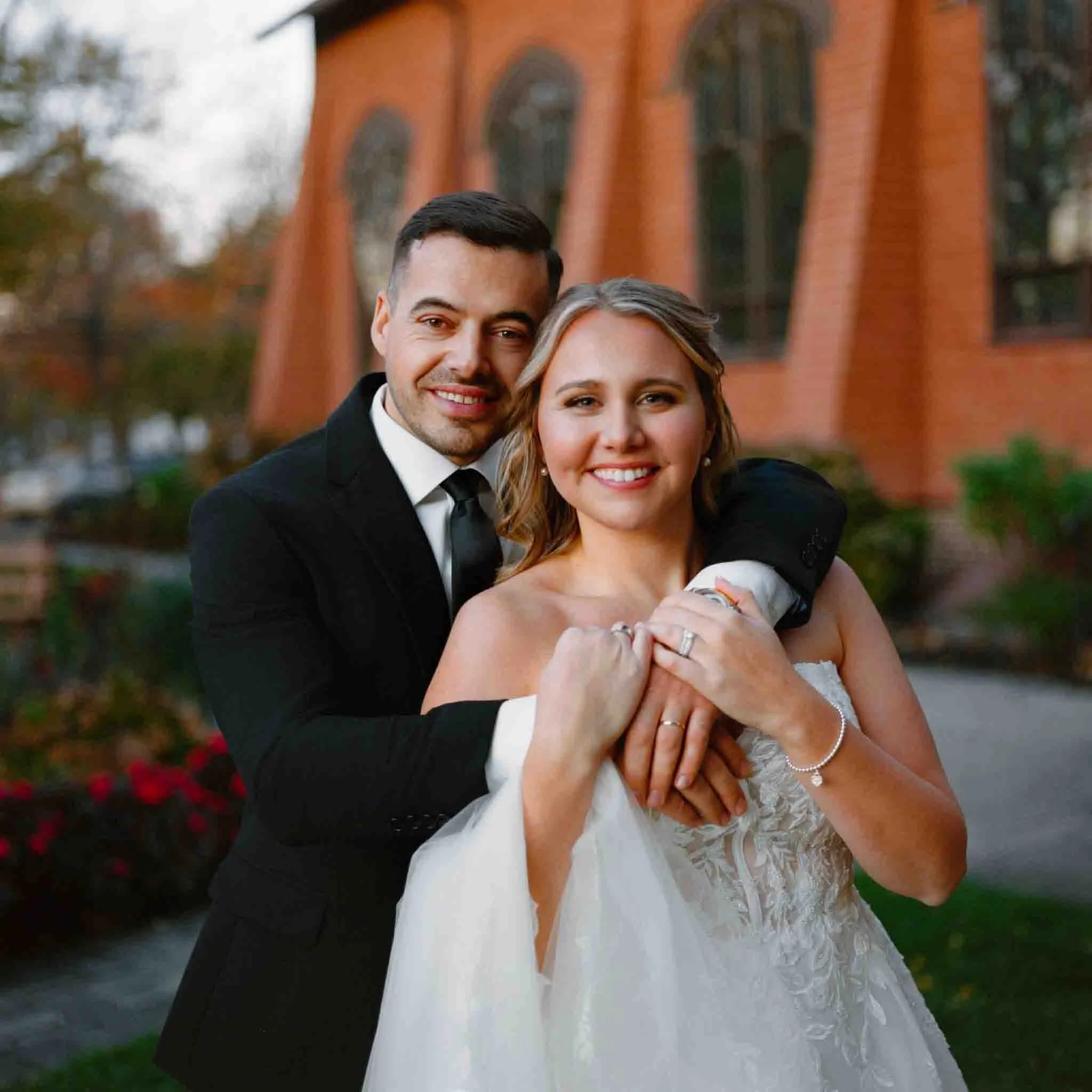 A smiling bride and groom pose outdoors in front of a red brick building with arched windows. Captured by a Documentary wedding photographer, the groom wears a black suit, and the bride in white holds his hands over her chest.