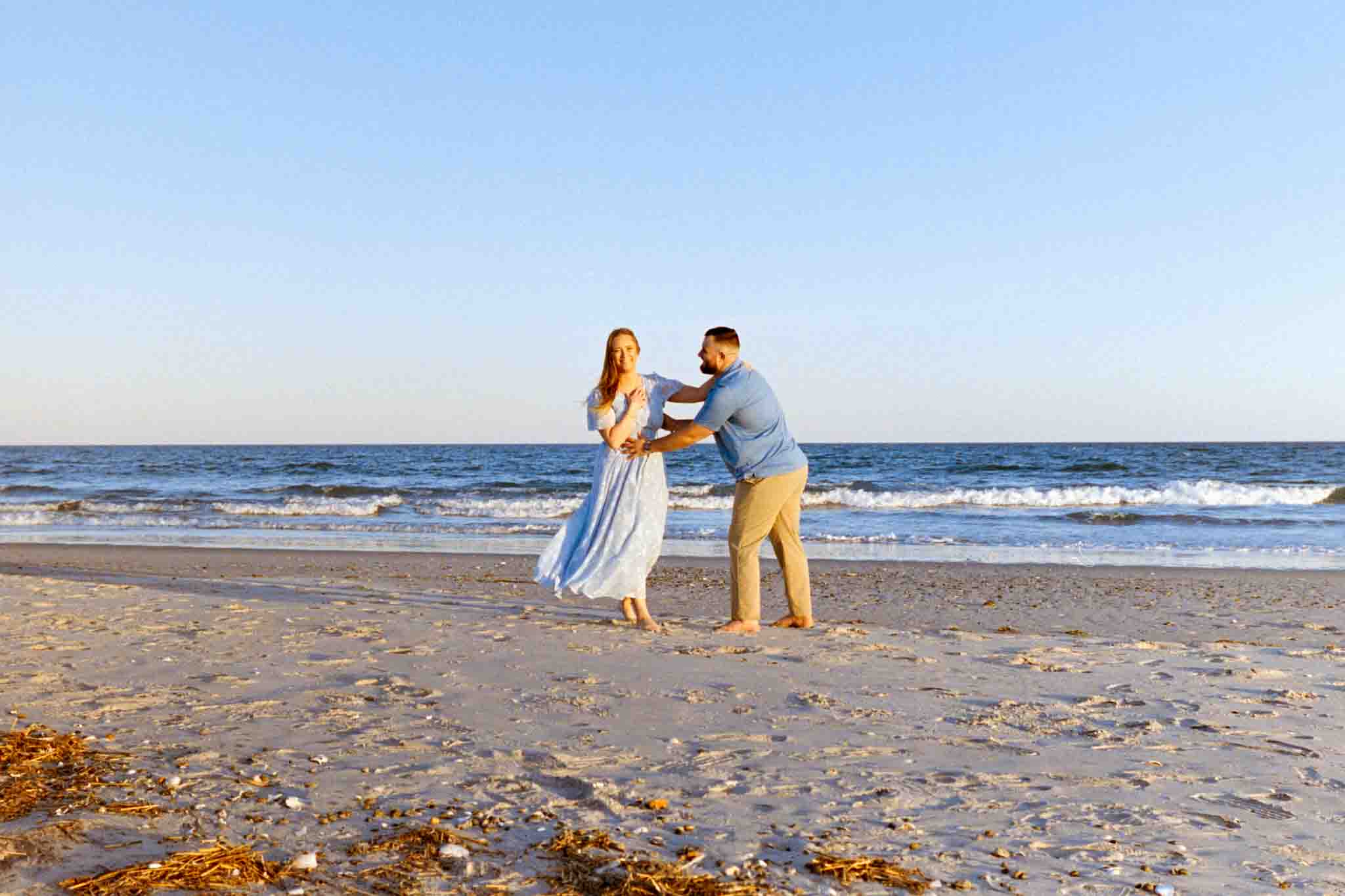 A woman in a light blue dress and a man in a blue shirt and khaki pants playfully spin together on a sandy beach with gentle waves, perfectly captured by a film wedding photographer NJ.