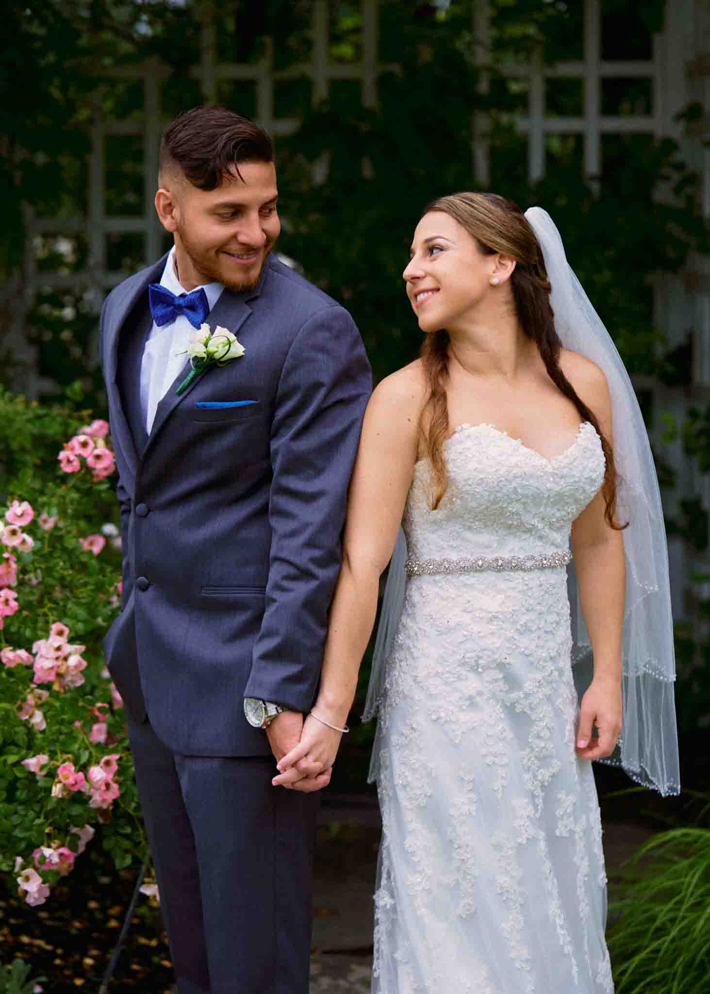 A bride and groom stand outdoors, holding hands and smiling at each other. Captured by a documentary wedding photographer, the groom wears a blue suit with a bow tie, while the bride wears a white lace gown. Pink flowers and greenery surround them.
