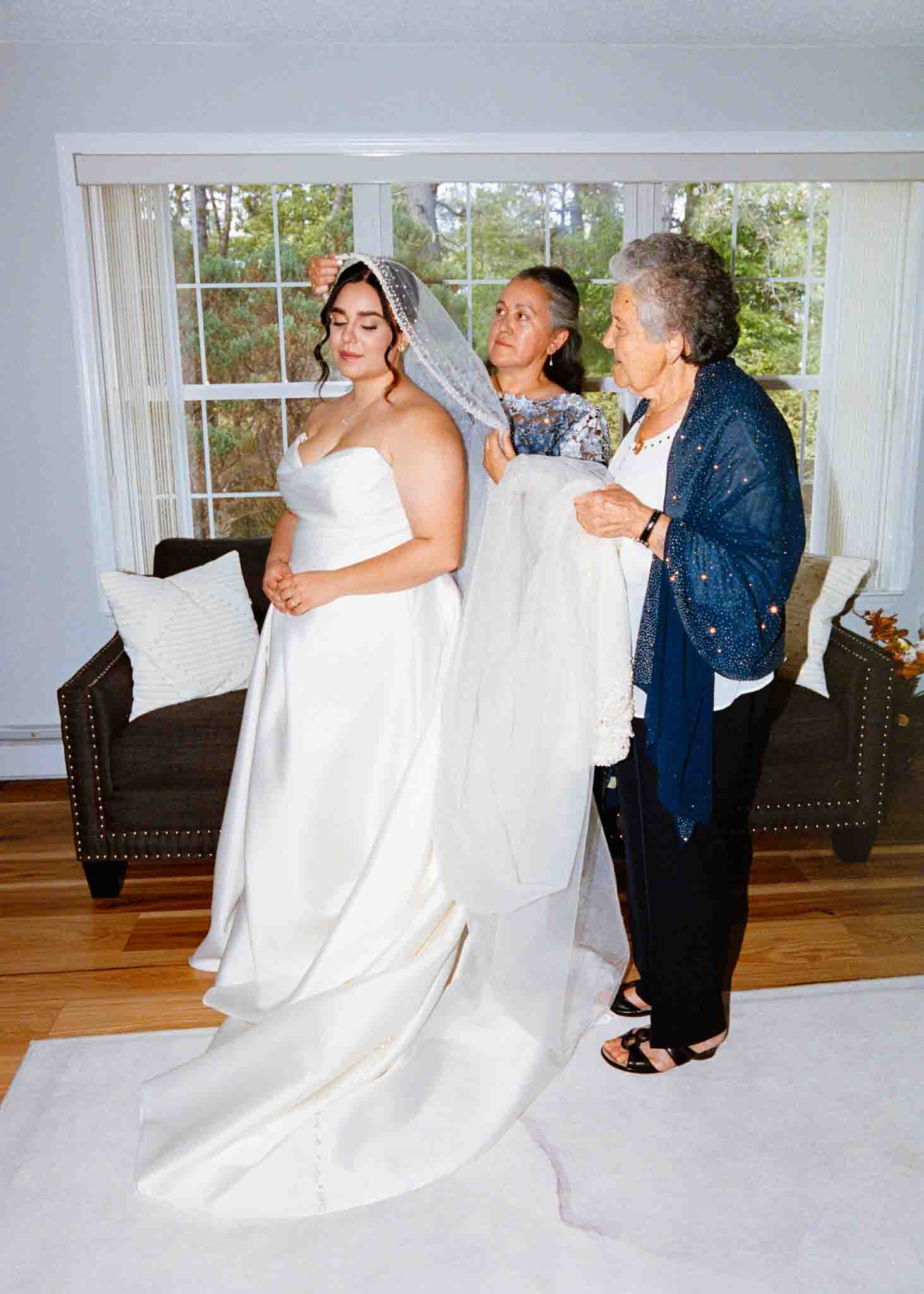 A bride in a white gown stands as two women help adjust her veil. Indoors by large windows with trees outside, the scene captures a warm, intimate moment—perfect for candid film wedding photos by a New Jersey documentary wedding photographer.