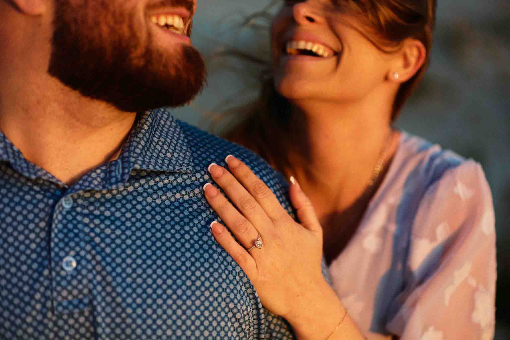 A close-up of a smiling couple, captured in warm, natural light. The woman rests her left hand, showing an engagement ring, on the mans shoulder. Perfectly documenting love through New Jersey film wedding photography.