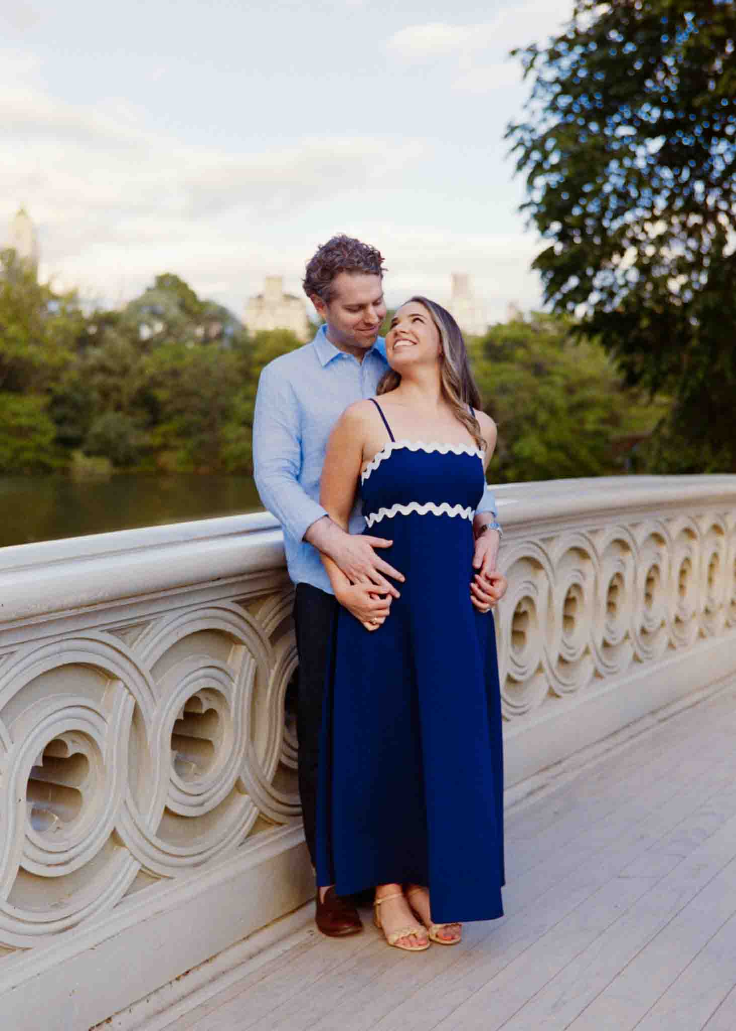 A couple stands on a decorative bridge, the man embracing the woman in a blue dress as she smiles up at him. Surrounded by greenery and a cloudy sky, this candid film wedding photo captures authentic moments by a New Jersey film wedding photography expert.