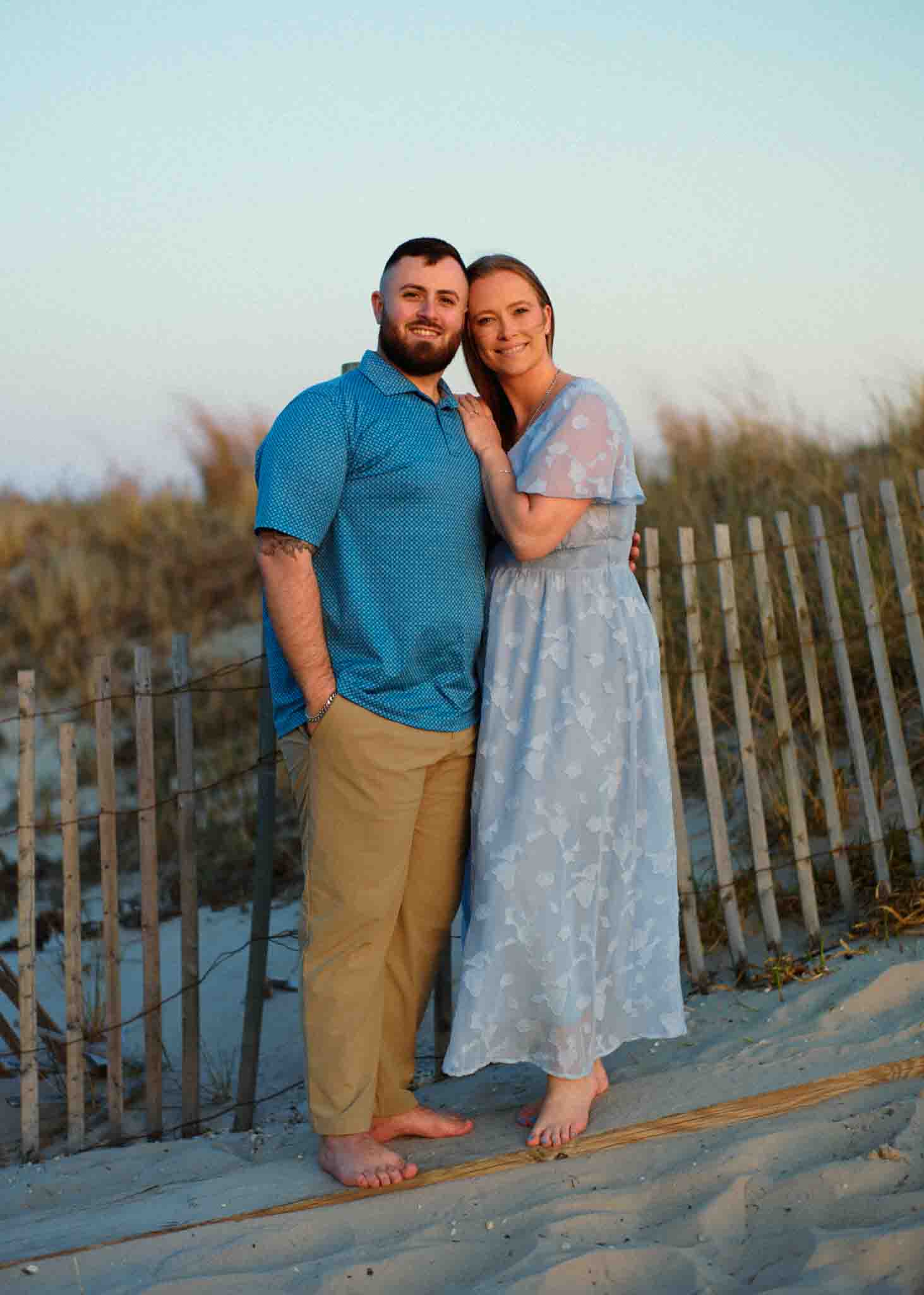 A barefoot man and woman stand close together on a sandy beach at sunset, smiling. The man wears a blue shirt and tan pants; the woman wears a light blue floral dress. Captured by a New Jersey film wedding photographer, beach grass and a wooden fence are in the background.