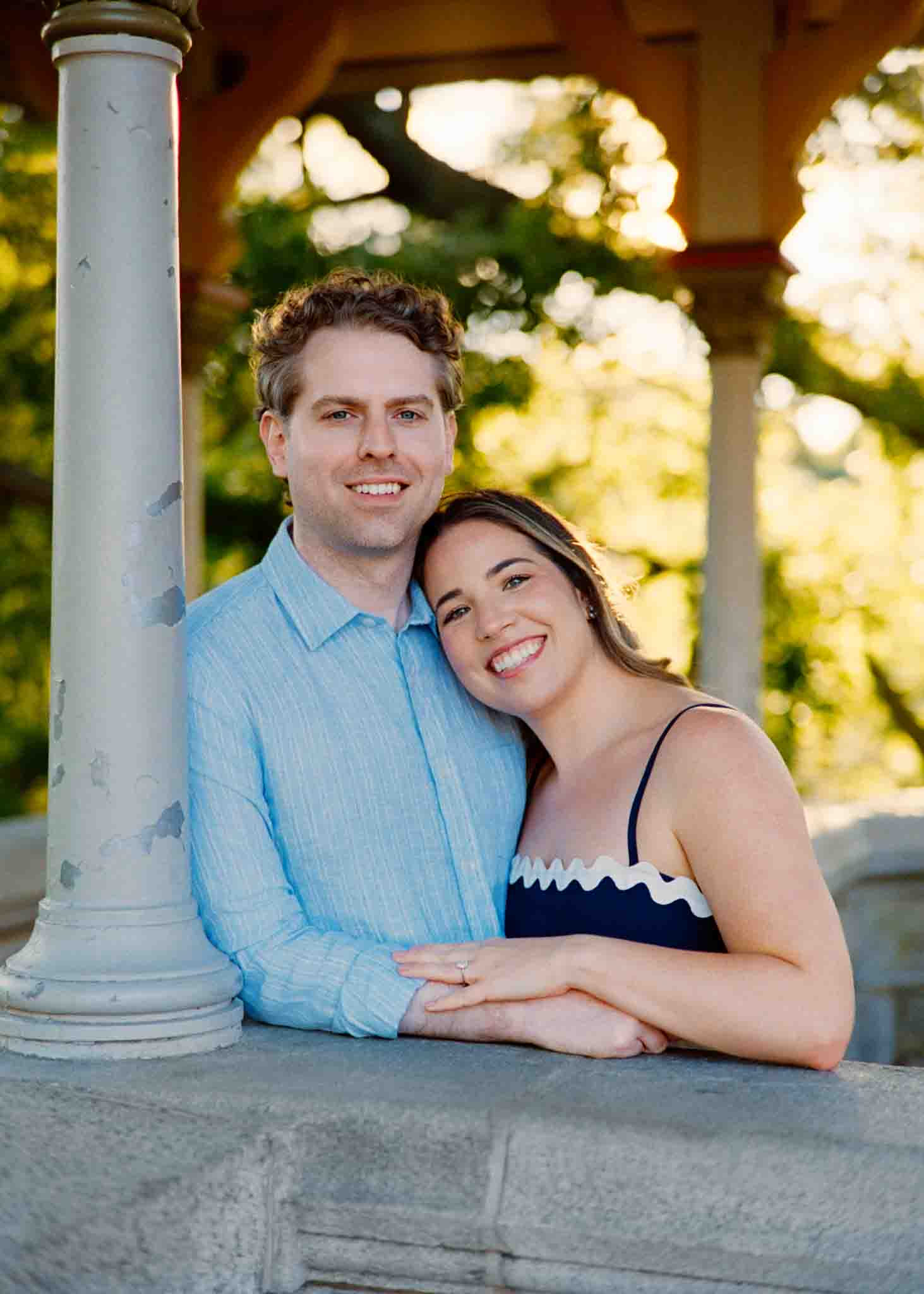 A smiling couple poses together outdoors, leaning against a stone railing under a gazebo. Captured by a film wedding photographer NJ, the woman rests her head on the mans shoulder as they enjoy greenery and sunlight in the background.