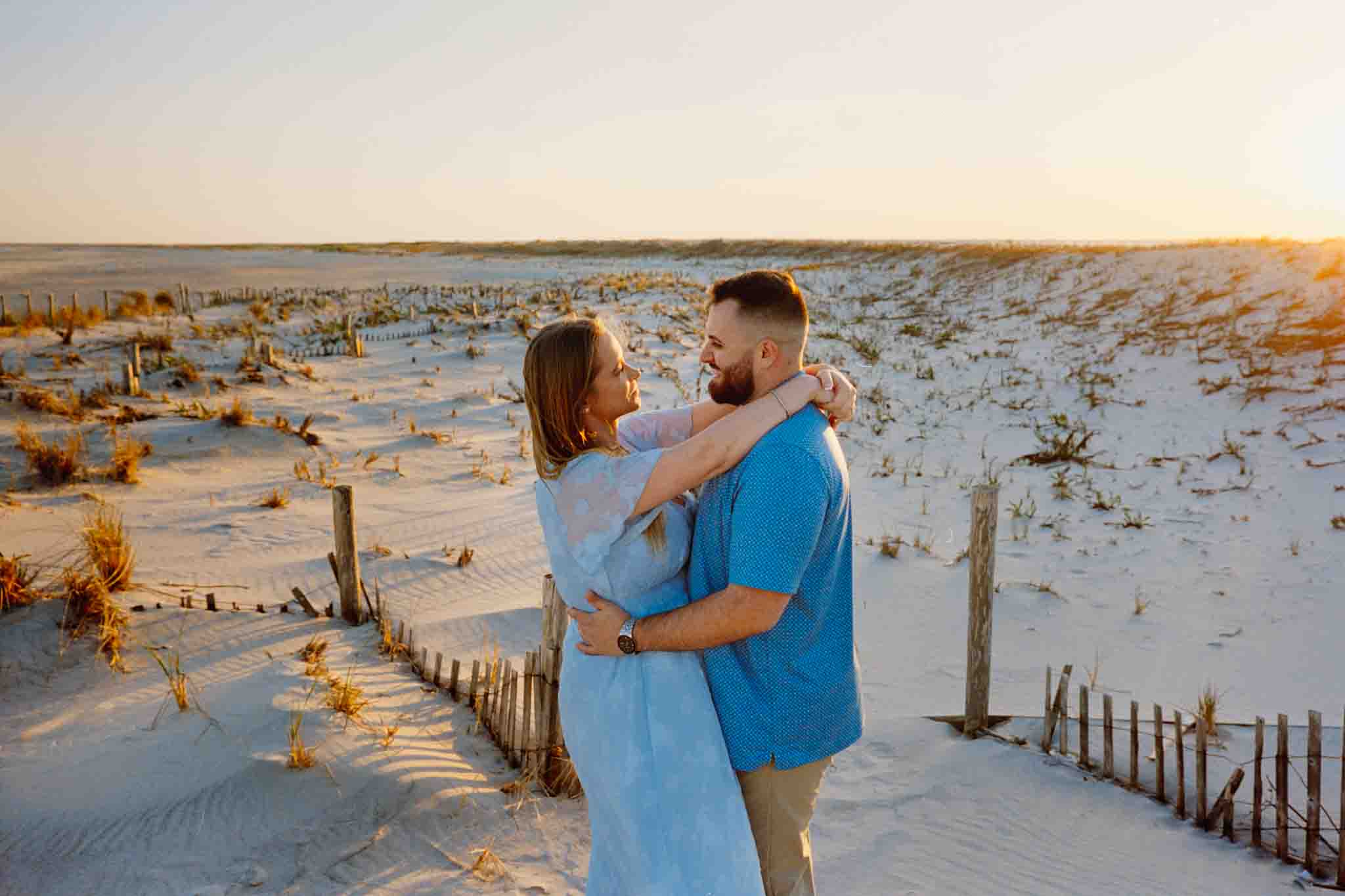 A couple embraces and smiles at each other on a sandy beach at sunset, with dunes, dried grasses, and a wooden fence in the background—perfect for candid film wedding photos New Jersey couples will cherish.