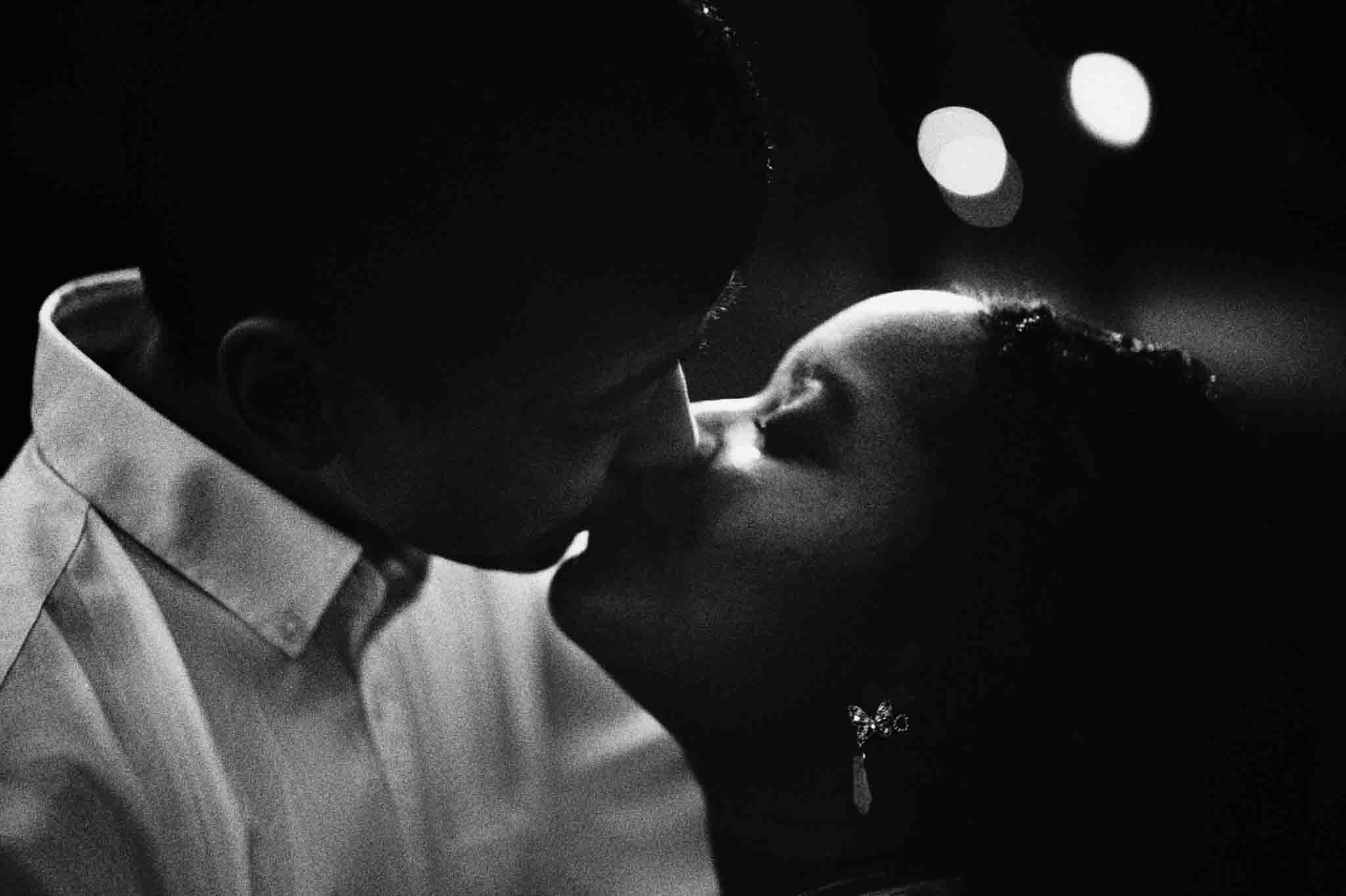 A black and white close-up of a couple sharing a kiss, captured by an analog wedding photography NJ artist. The man wears a collared shirt, the woman has curly hair and an earring, with softly blurred circular light spots in the background.