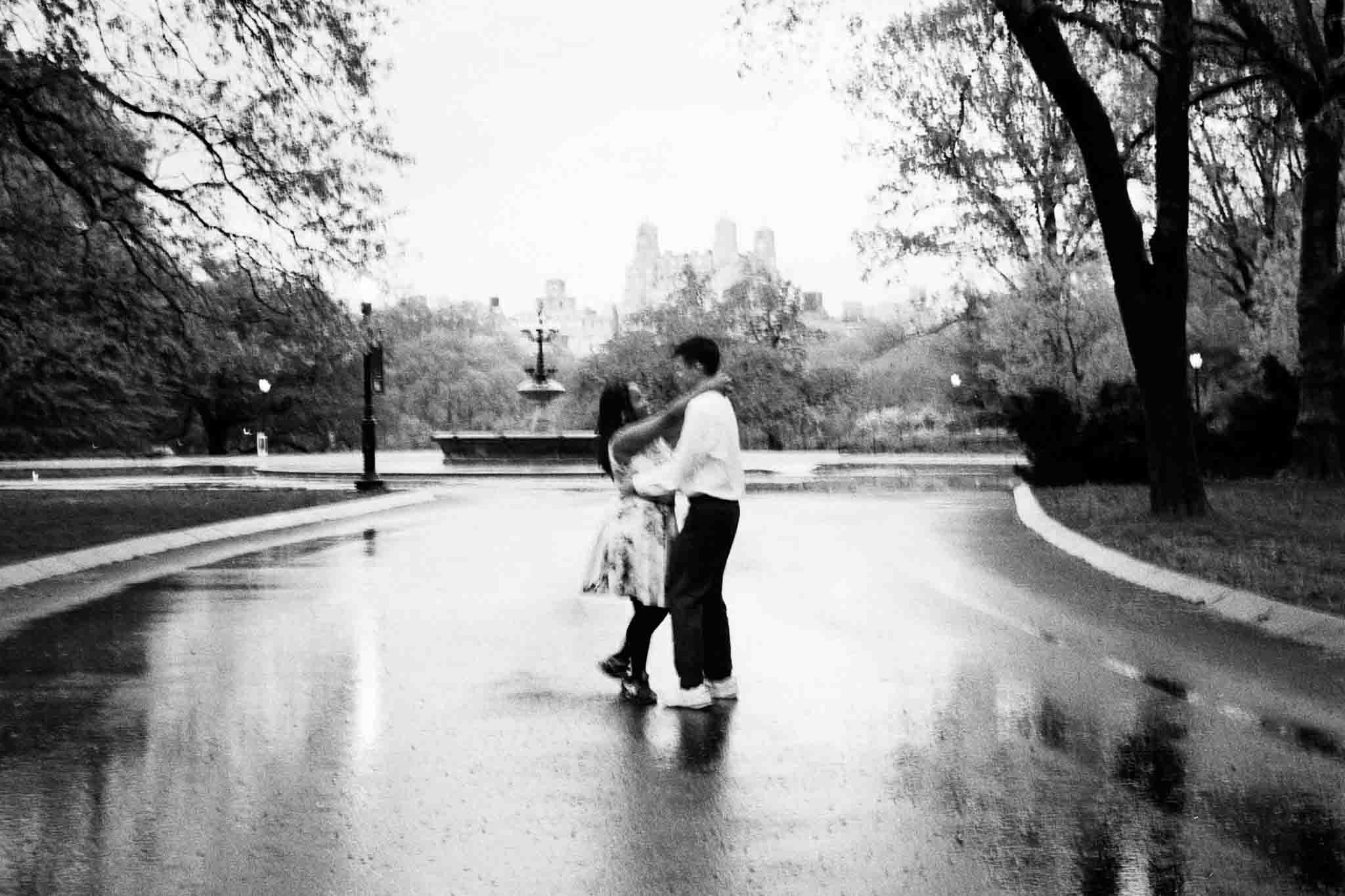 A couple dances together on a wet, empty park path on a rainy day, surrounded by trees and distant blurred buildings—a scene reminiscent of candid film wedding photos New Jersey couples cherish.