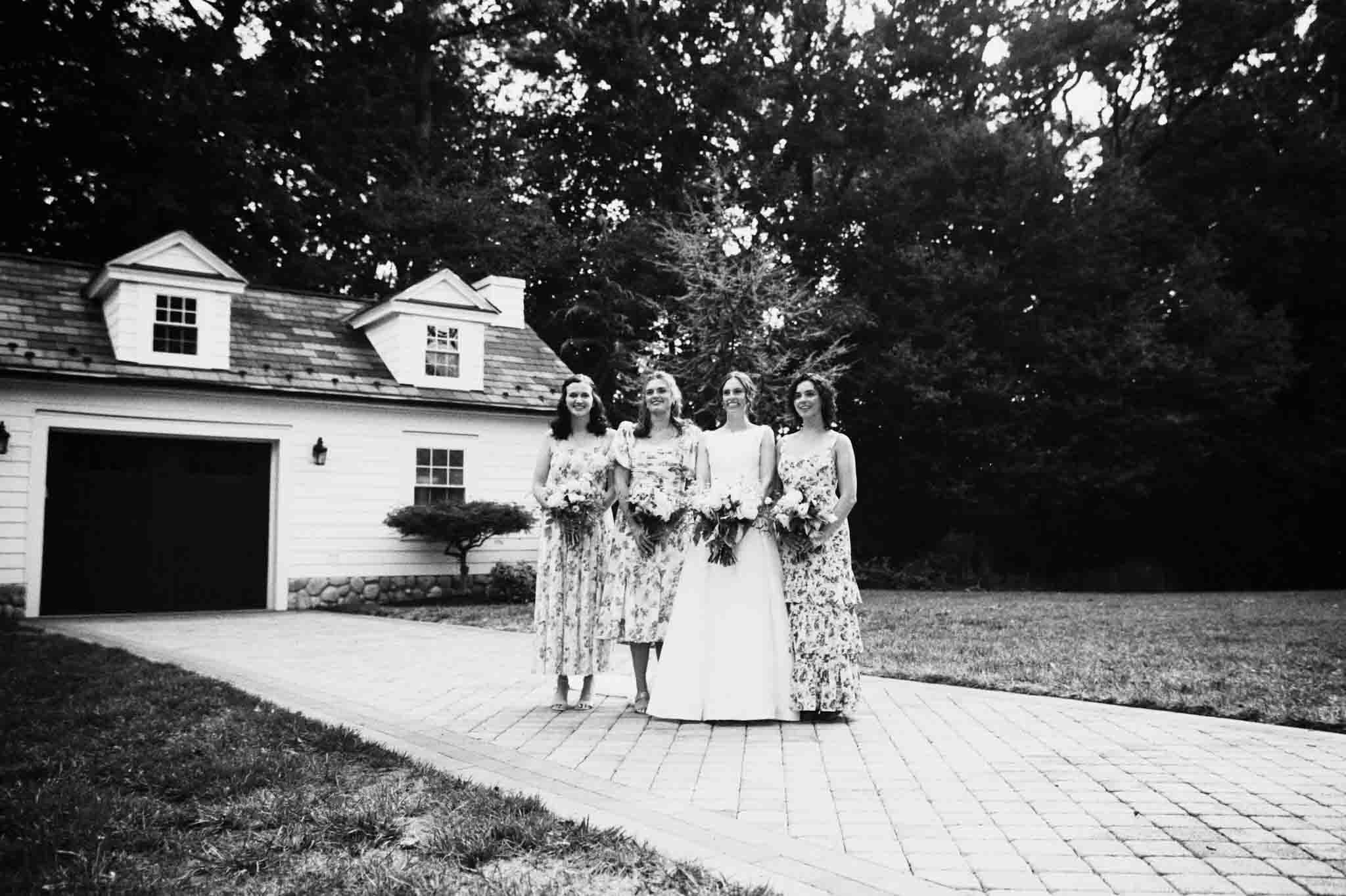 Four women in dresses, one in a bridal gown, smile and hold bouquets on a paved driveway before a white house with a garage and trees. This black and white image captures the charm of candid film wedding photos New Jersey couples love.