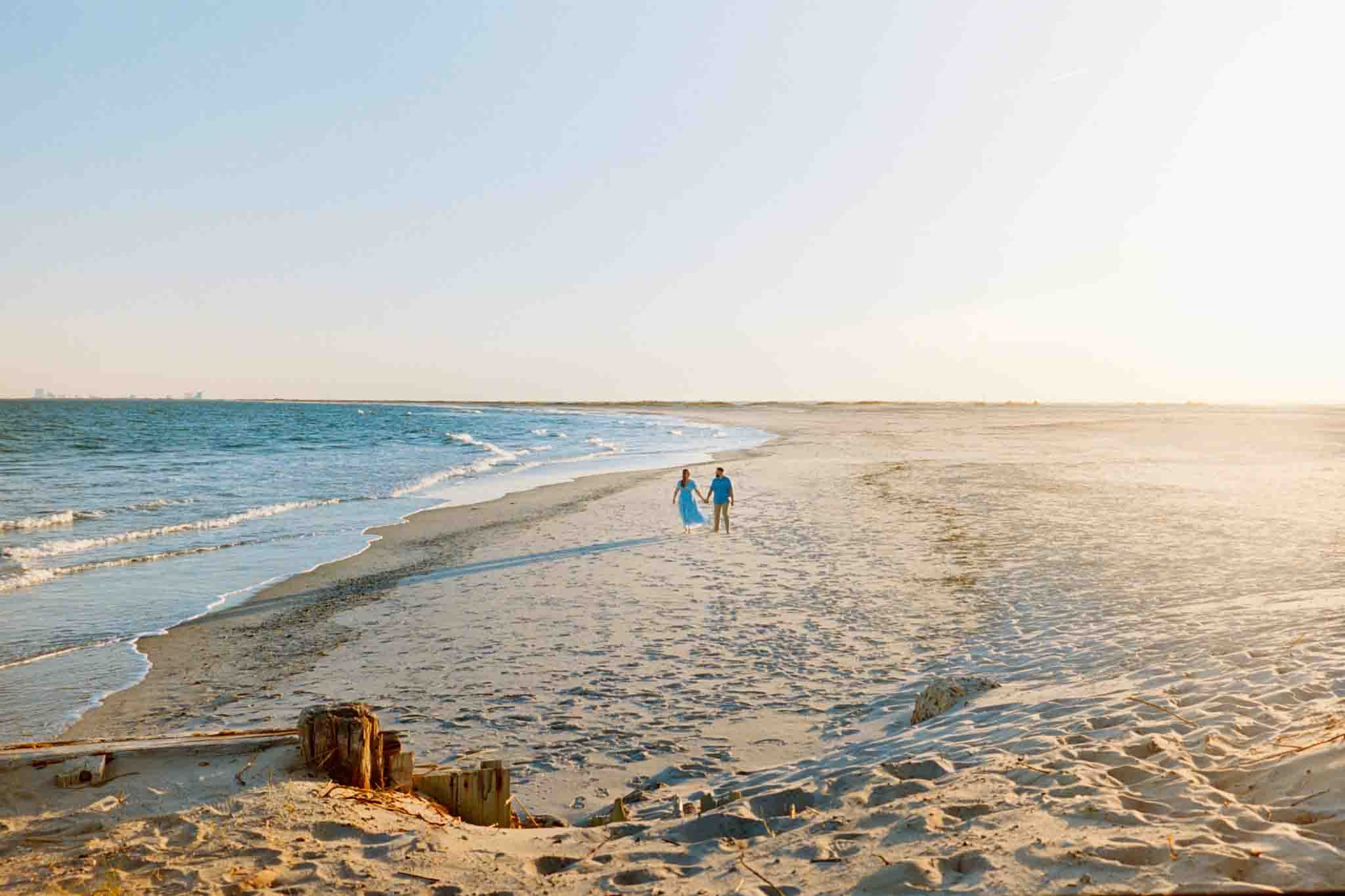Two people walk hand in hand along a sandy beach at sunset, with gentle waves on one side and a clear sky above. Captured by a documentary wedding photographer, the scene evokes the candid film wedding photos New Jersey couples cherish.