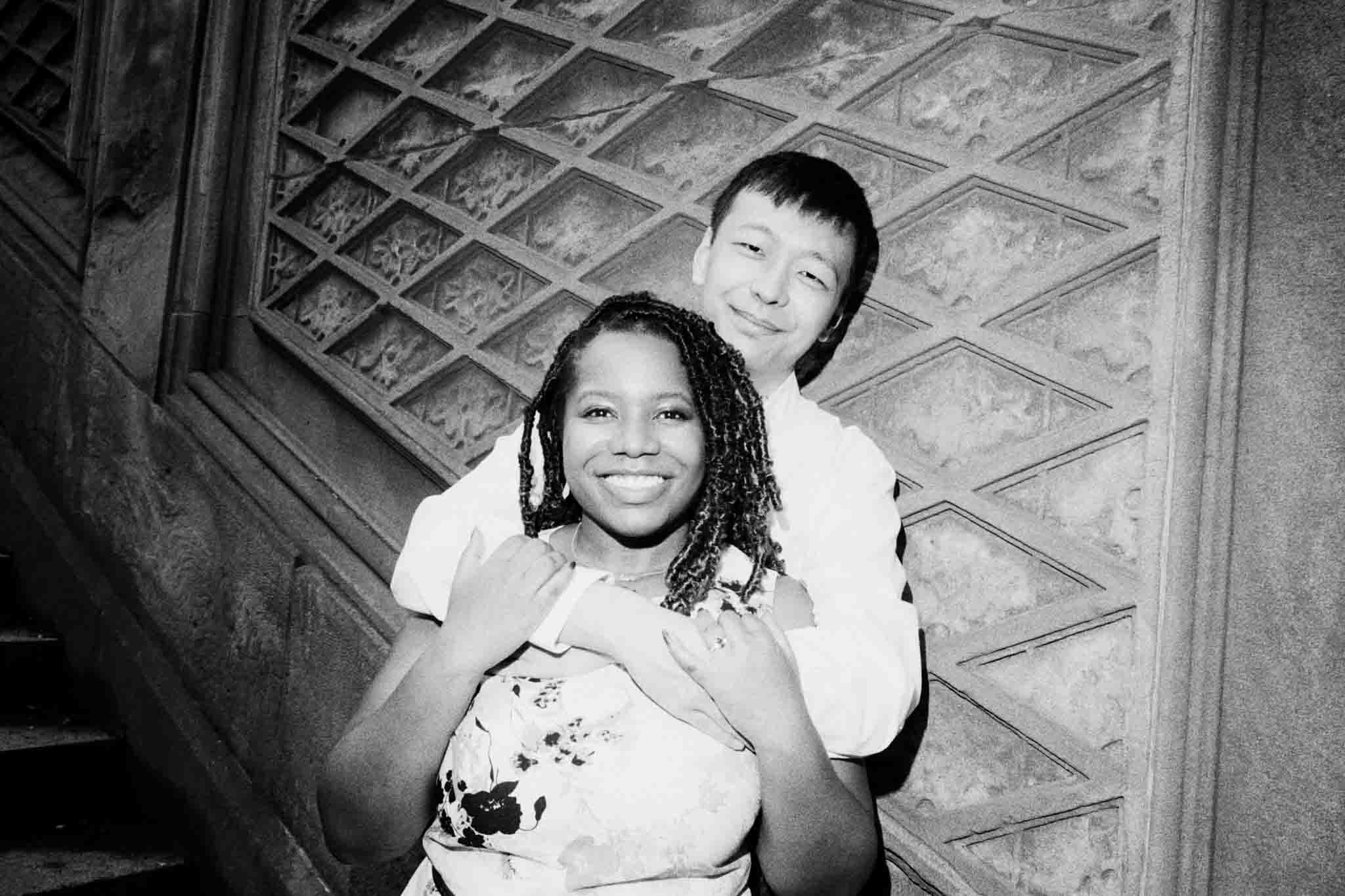 A smiling couple poses closely together in front of an ornate, patterned wall. Captured by a documentary wedding photographer, the woman stands in front holding the man’s arms as he hugs her from behind; both look genuinely happy.