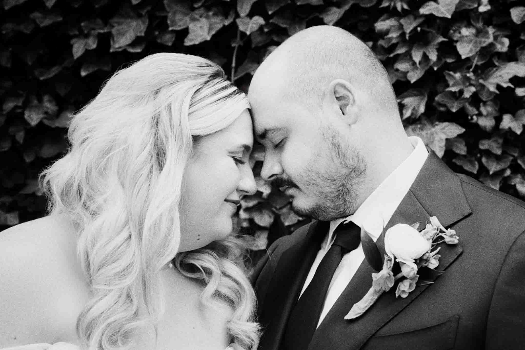 A couple in wedding attire gently touch foreheads and smile with their eyes closed, surrounded by greenery—captured in candid film wedding photos New Jersey.