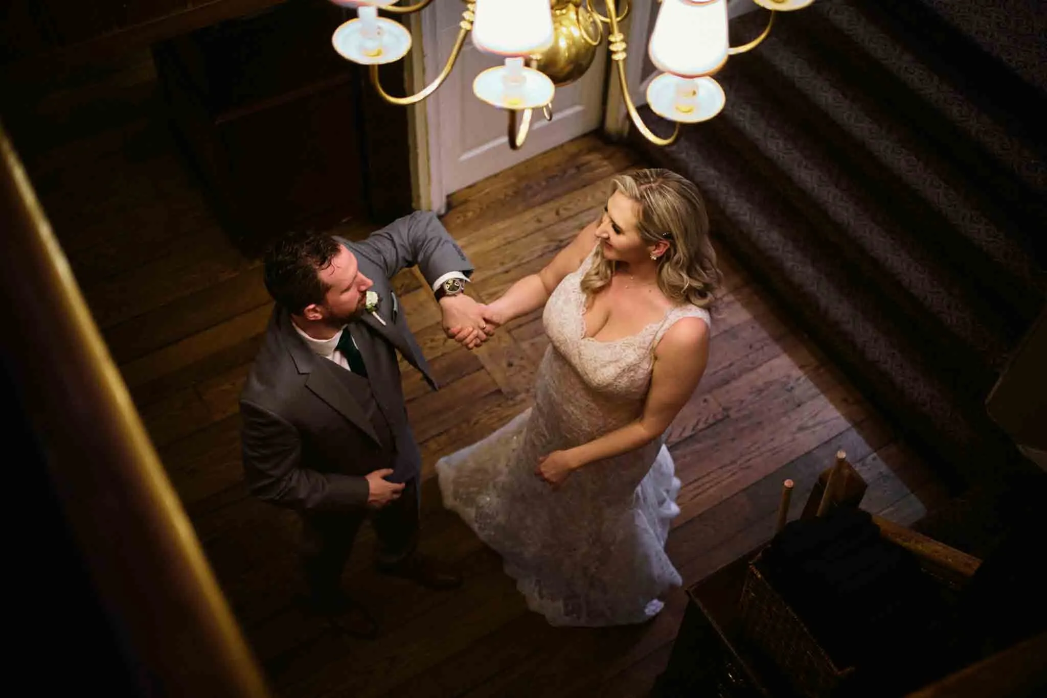 A bride and groom, seen from above, hold hands and smile at each other while standing on a wooden floor near a staircase and under a chandelier, beautifully captured by an analog wedding photography NJ expert.