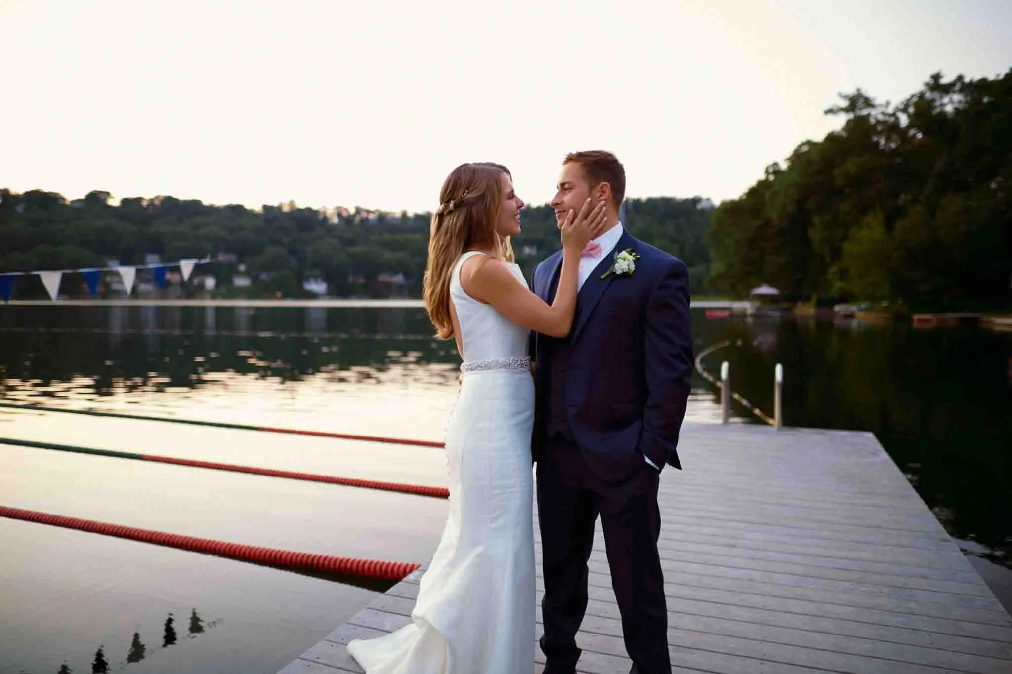 A bride in a white dress and a groom in a dark suit stand on a wooden dock by a calm lake, gazing at each other lovingly, as candid film wedding photos New Jersey capture the moment among trees and distant cabins.