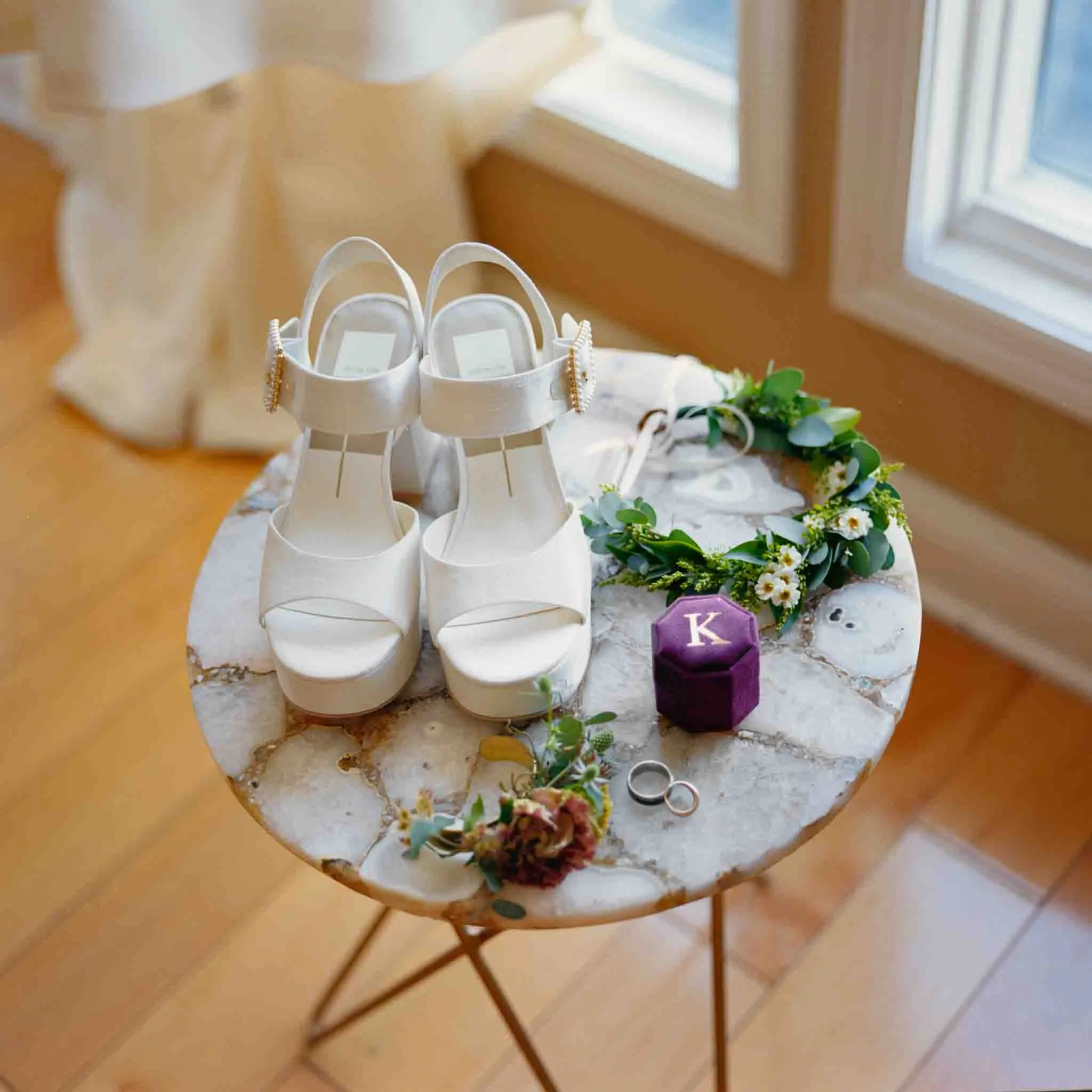 White platform bridal shoes, wedding rings, a floral boutonniere, a purple ring box with the letter K, and a greenery headpiece are beautifully arranged on a small round marble table by a sunny window—perfect for a documentary wedding photographer.
