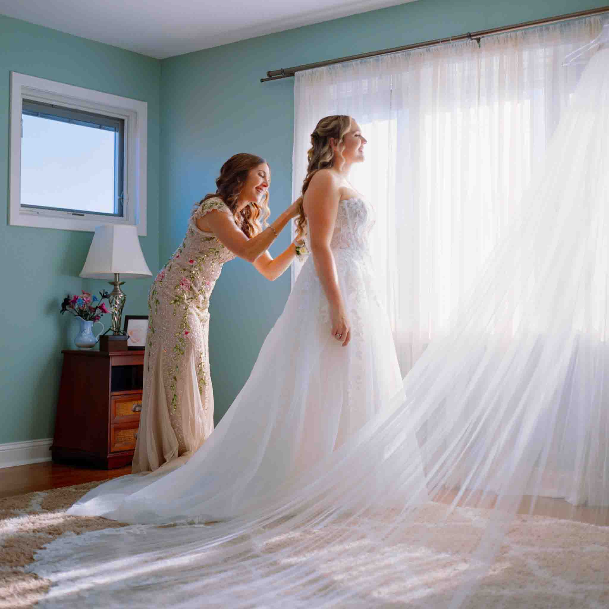 A woman helps a bride adjust her strapless white wedding gown in a sunlit room with light blue walls and sheer curtains. Captured by a documentary wedding photographer, both women smile warmly as the bride’s long veil spreads out behind her.
