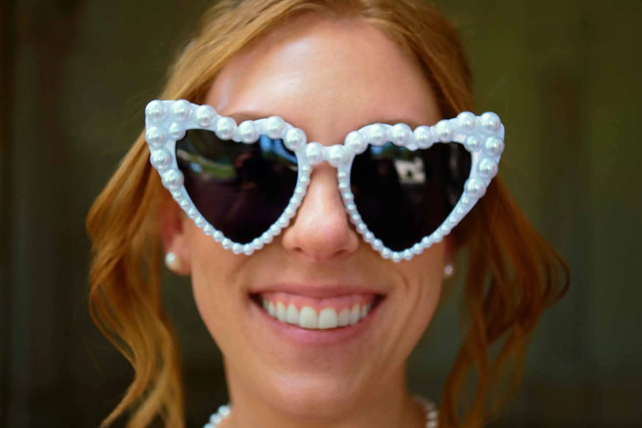 A smiling woman with wavy red hair wears large, heart-shaped sunglasses decorated with pearls. Captured in a style reminiscent of New Jersey film wedding photography, the blurred background brings focus to her face and accessories.