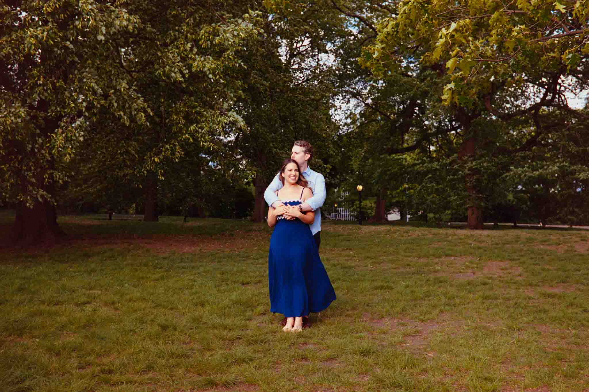 A couple stands together on grass in a park surrounded by green trees. Captured by a New Jersey film wedding photographer, the woman in a long blue dress smiles as the man embraces her from behind, his arms around her shoulders.