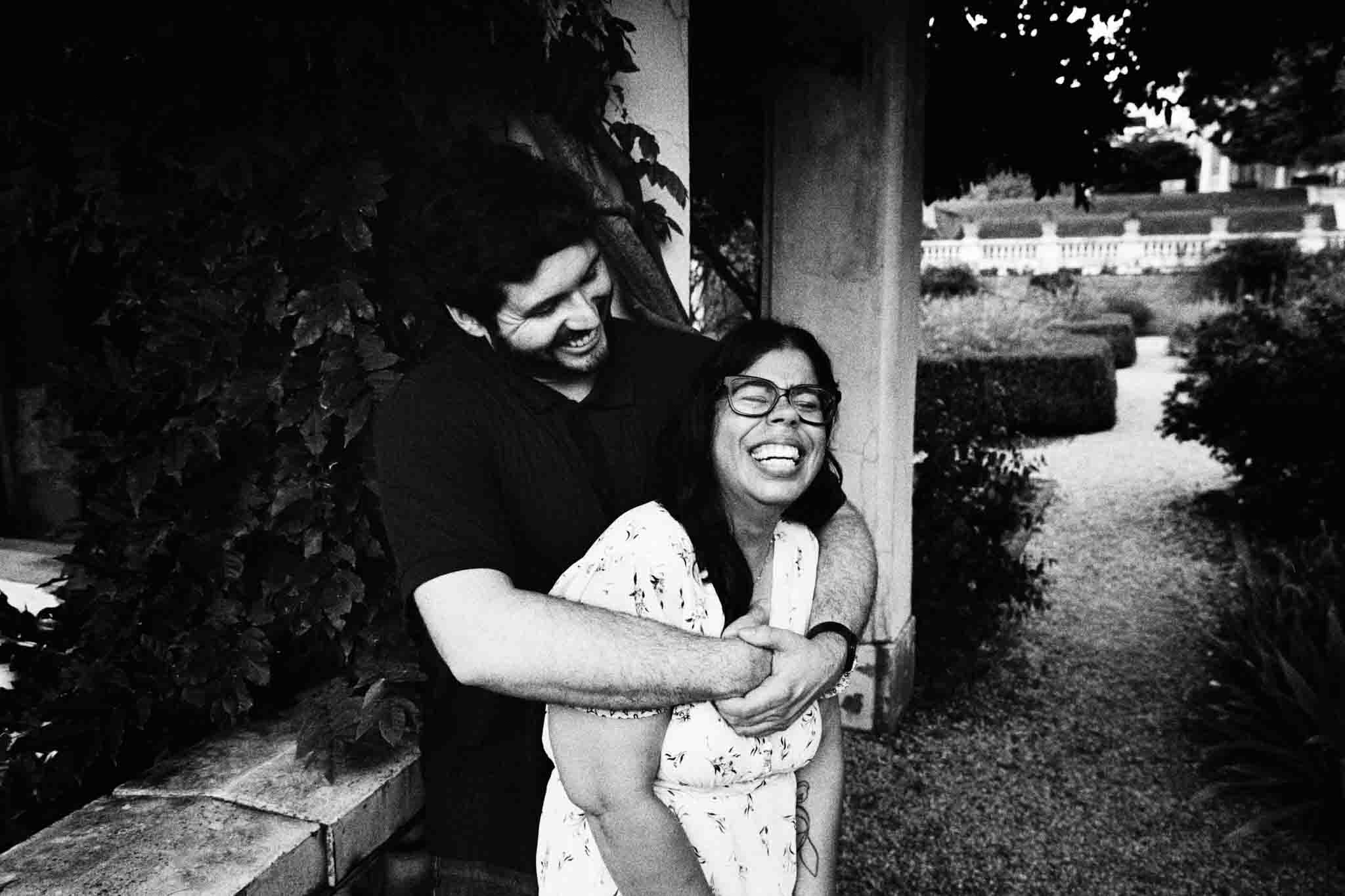 A man hugs a woman from behind as they both laugh joyfully in a garden setting. This candid film wedding photo, taken by a film wedding photographer NJ, features trees, bushes, and a building in the black and white background.