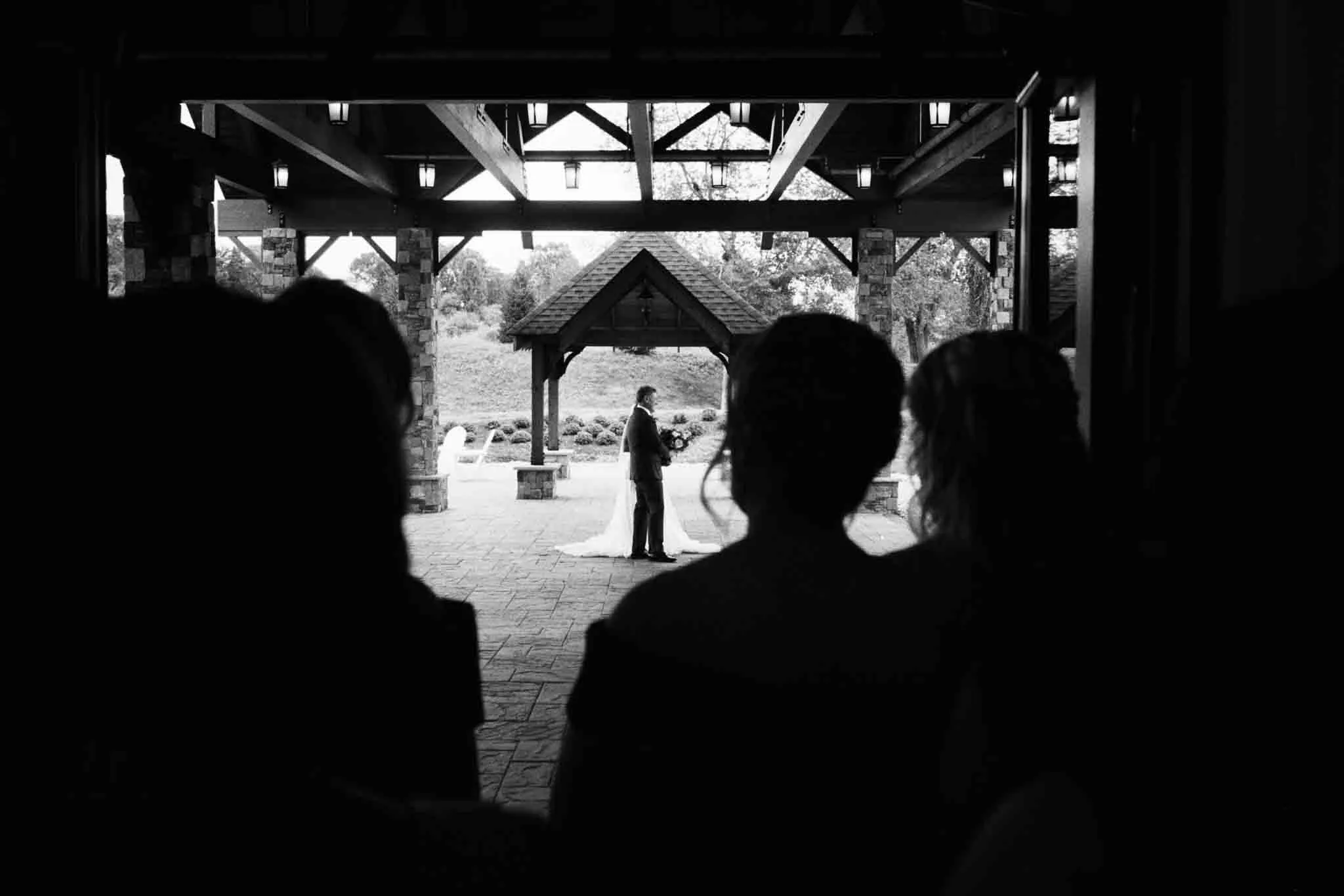 A bride and groom share a kiss under an outdoor pavilion, framed by silhouetted guests in the foreground—a candid film wedding photo by a documentary wedding photographer in New Jersey.